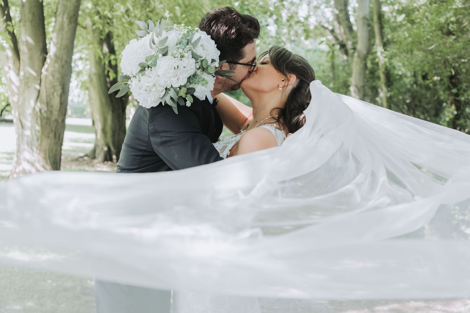 Bride and groom sharing a romantic kiss outdoors, with a flowing white veil and bridal bouquet of white flowers.