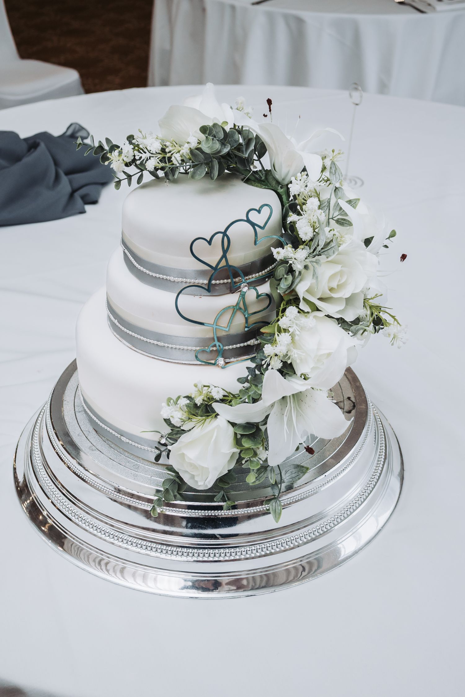 Elegant white wedding cake decorated with silver trim and fresh white roses on a white tablecloth.