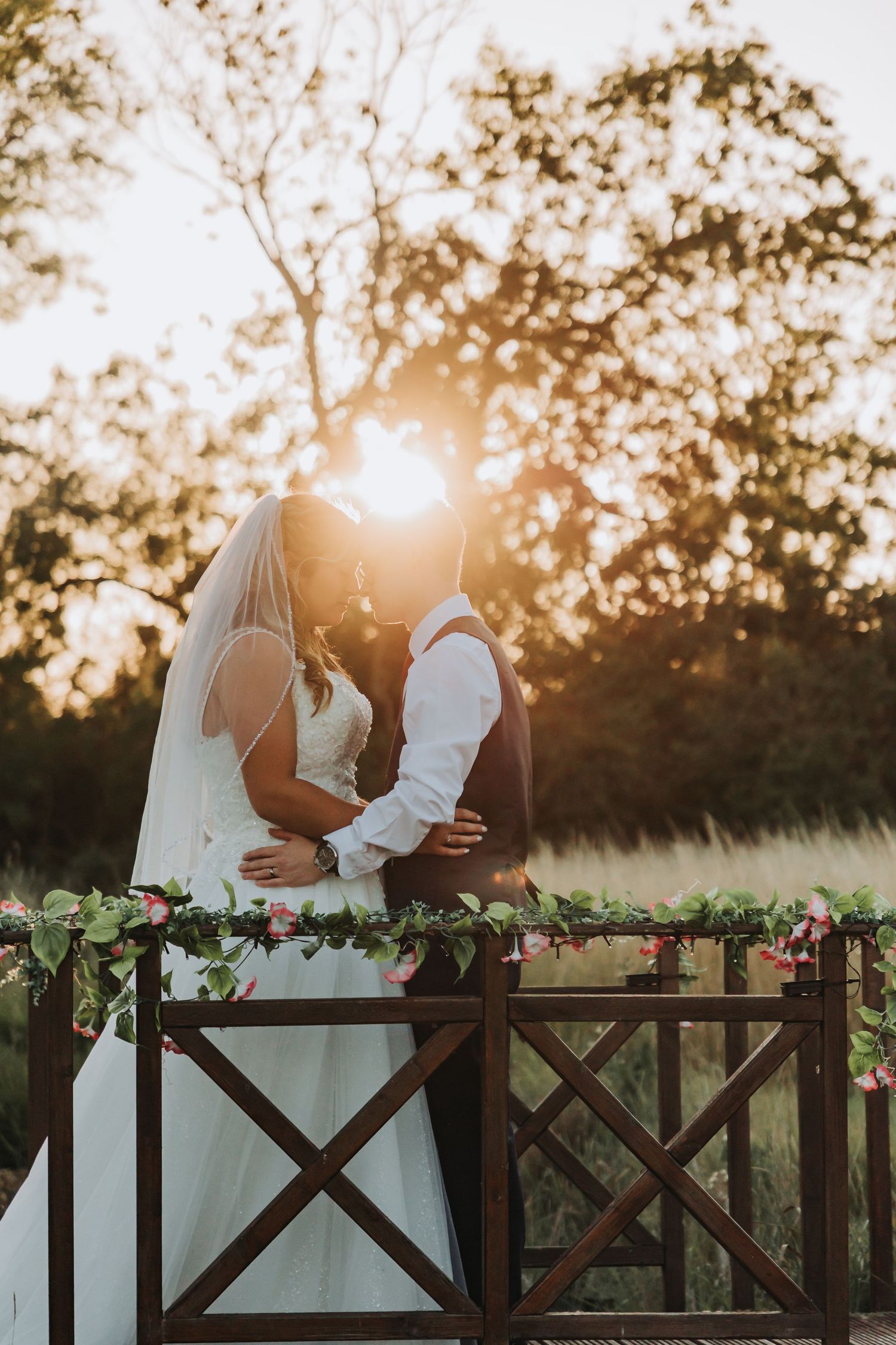 A couple in wedding attire sits together on a wooden fence at sunset with golden light streaming through trees.