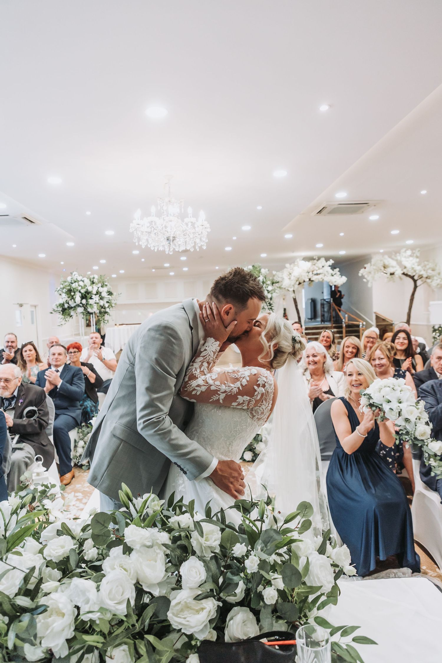 Wedding guests react with joy as newlyweds share their first kiss during an elegant indoor ceremony with floral decor.