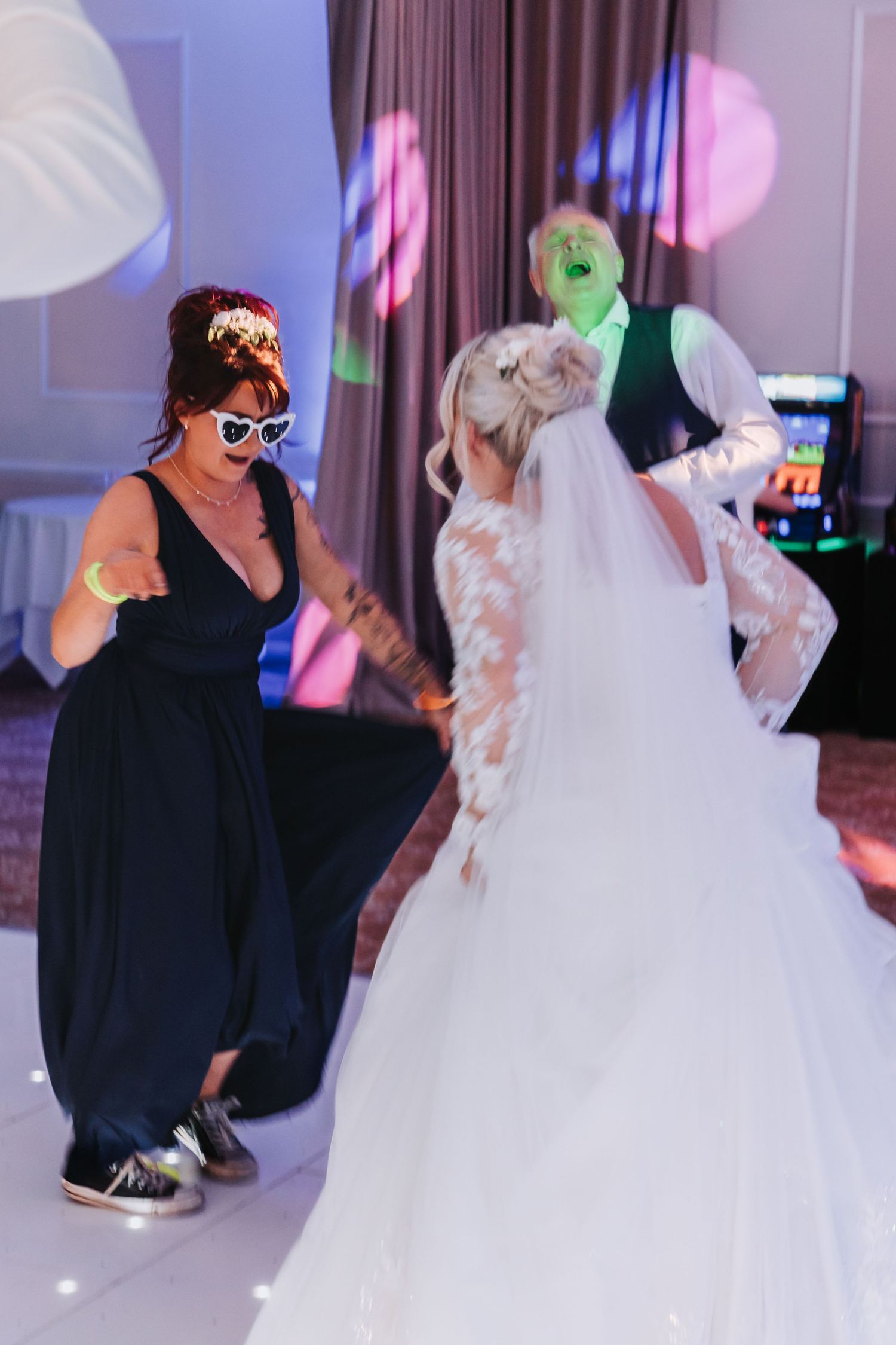 A bride in a white dress dances with someone wearing black at a wedding reception with colorful lighting.