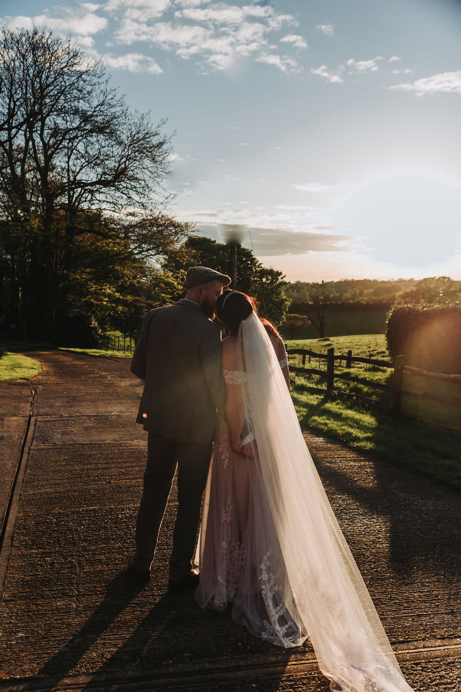 Silhouetted couple in wedding attire embrace during golden sunset on rustic path with wooden fence and trees.
