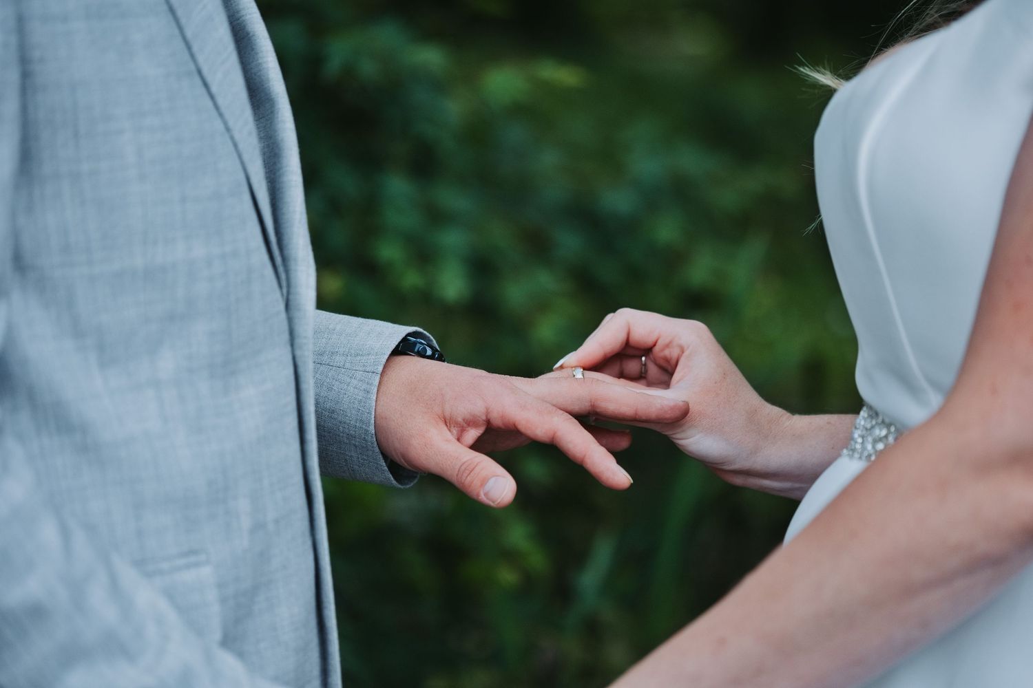 Bride placing a wedding ring on groom's finger during outdoor ceremony, with lush green foliage in background.