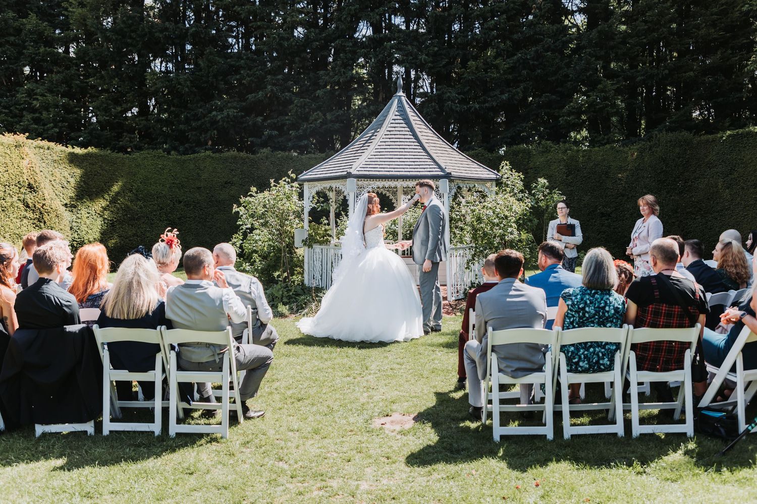 Outdoor garden wedding ceremony with white chairs and a decorative gazebo on a sunny afternoon.