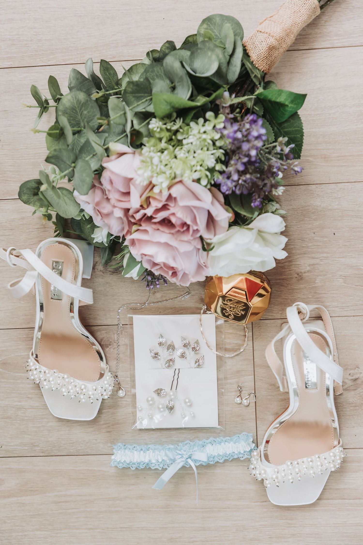 Wedding details flatlay with pink and white bouquet, pearl-embellished bridal shoes, blue garter and delicate jewelry on wood floor.