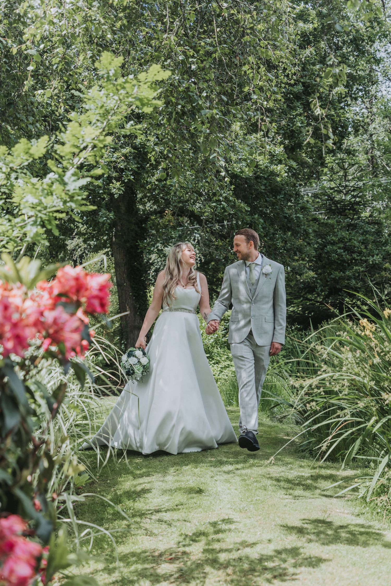 Bride and groom walk hand-in-hand through a lush garden path surrounded by green trees and pink flowers.