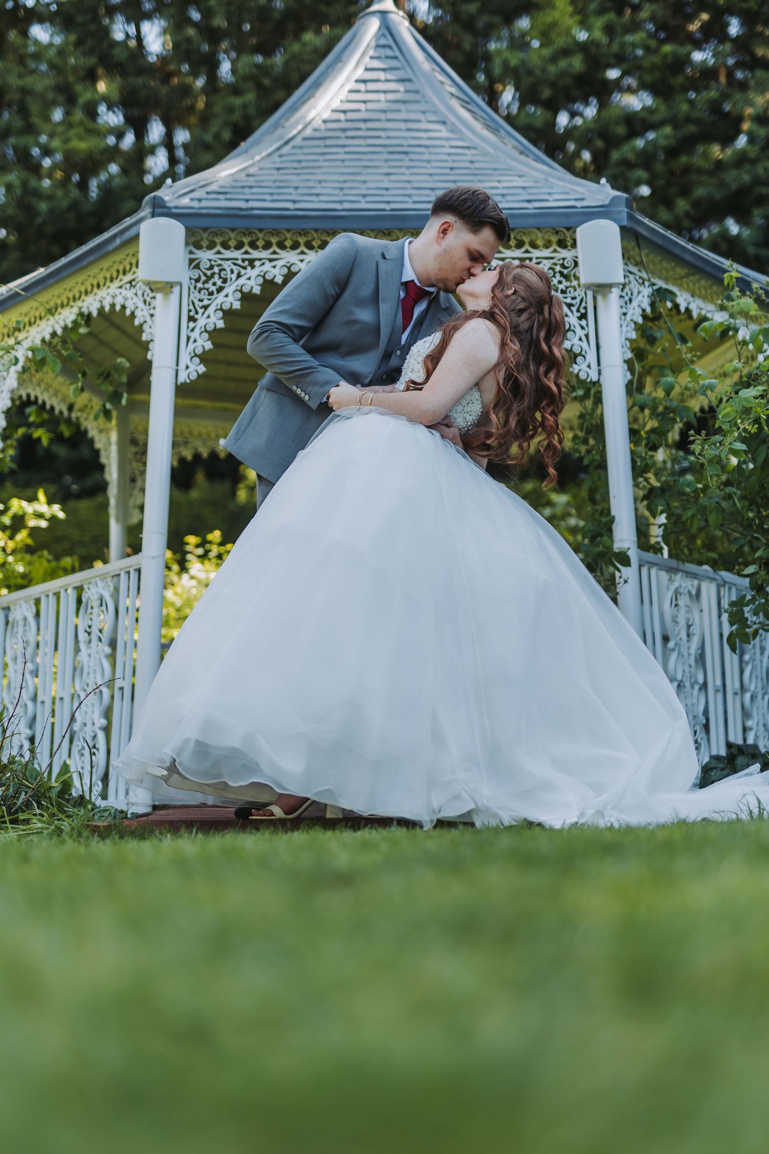 A couple poses together under a white wooden gazebo surrounded by greenery during their wedding day.