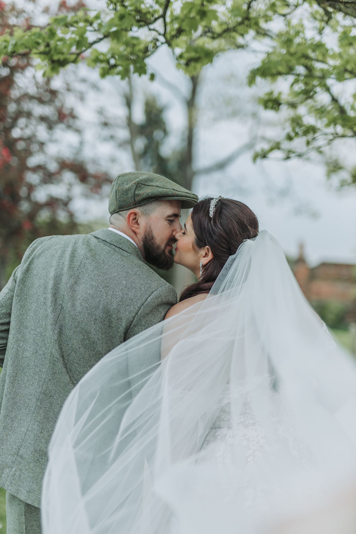 Romantic wedding couple shares tender kisses under green spring foliage in an outdoor setting.