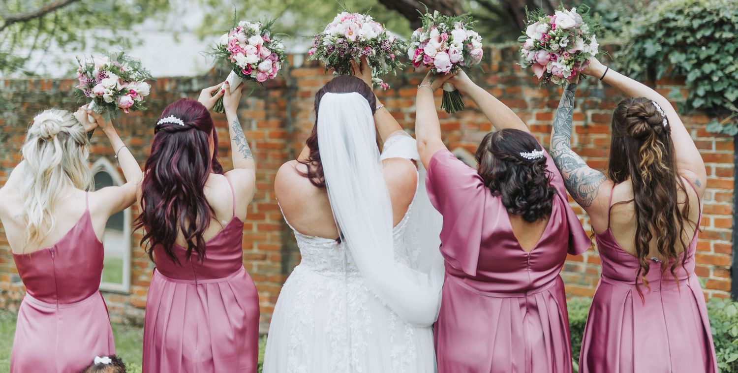 Bridesmaids in dusty rose dresses walk with bride in white gown along brick wall and garden path.
