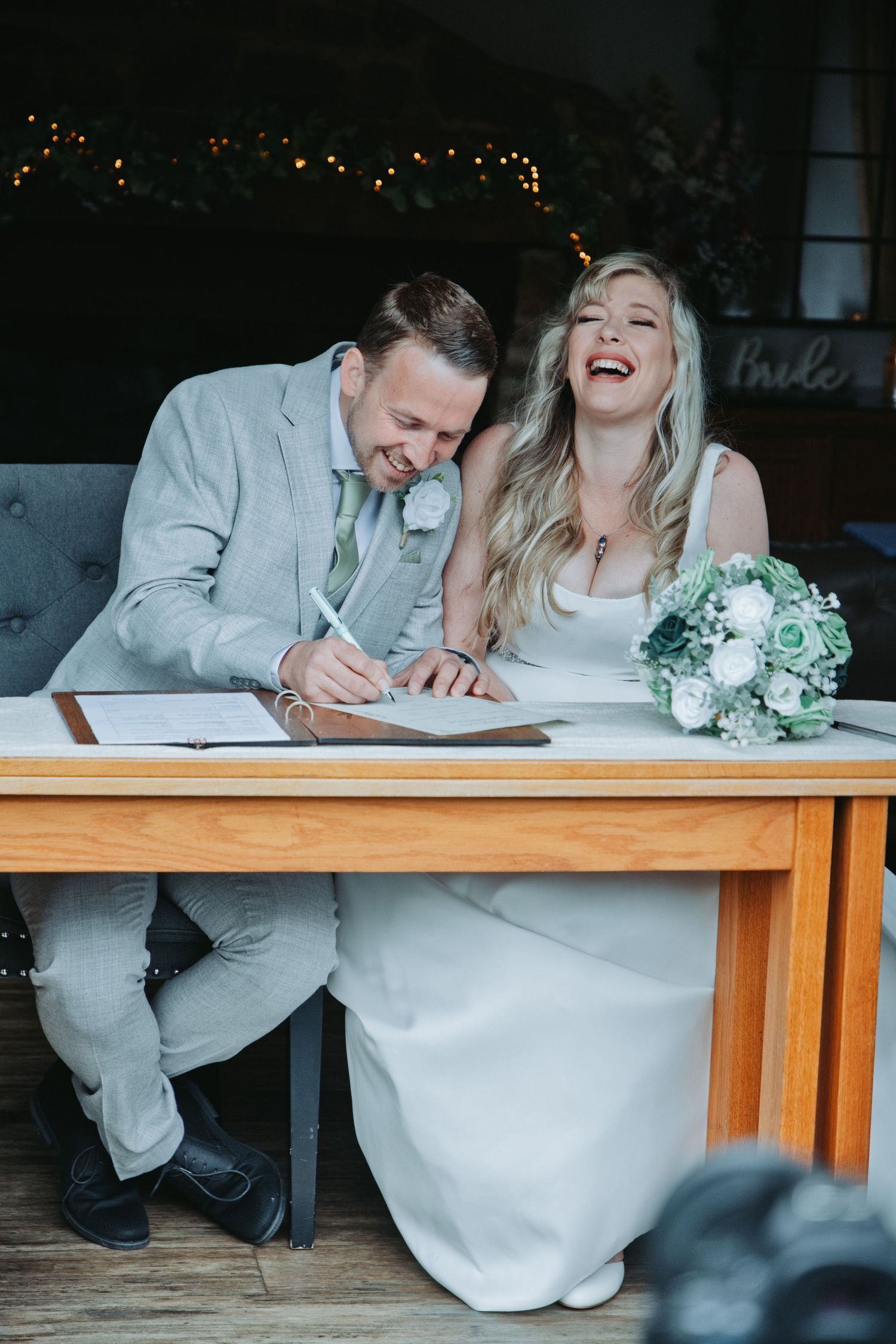 Laughing bride and groom signing wedding register at wooden table, bride holding white and green bouquet.