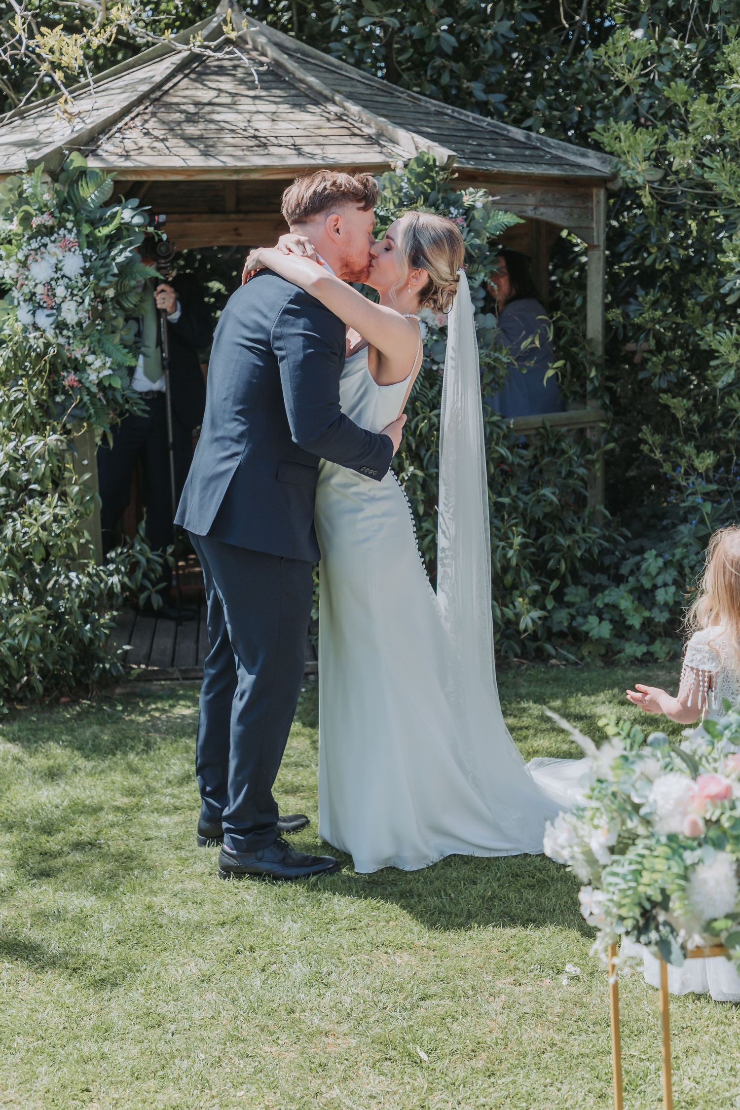 A couple shares a romantic kiss under a garden gazebo decorated with floral arrangements on their wedding day.