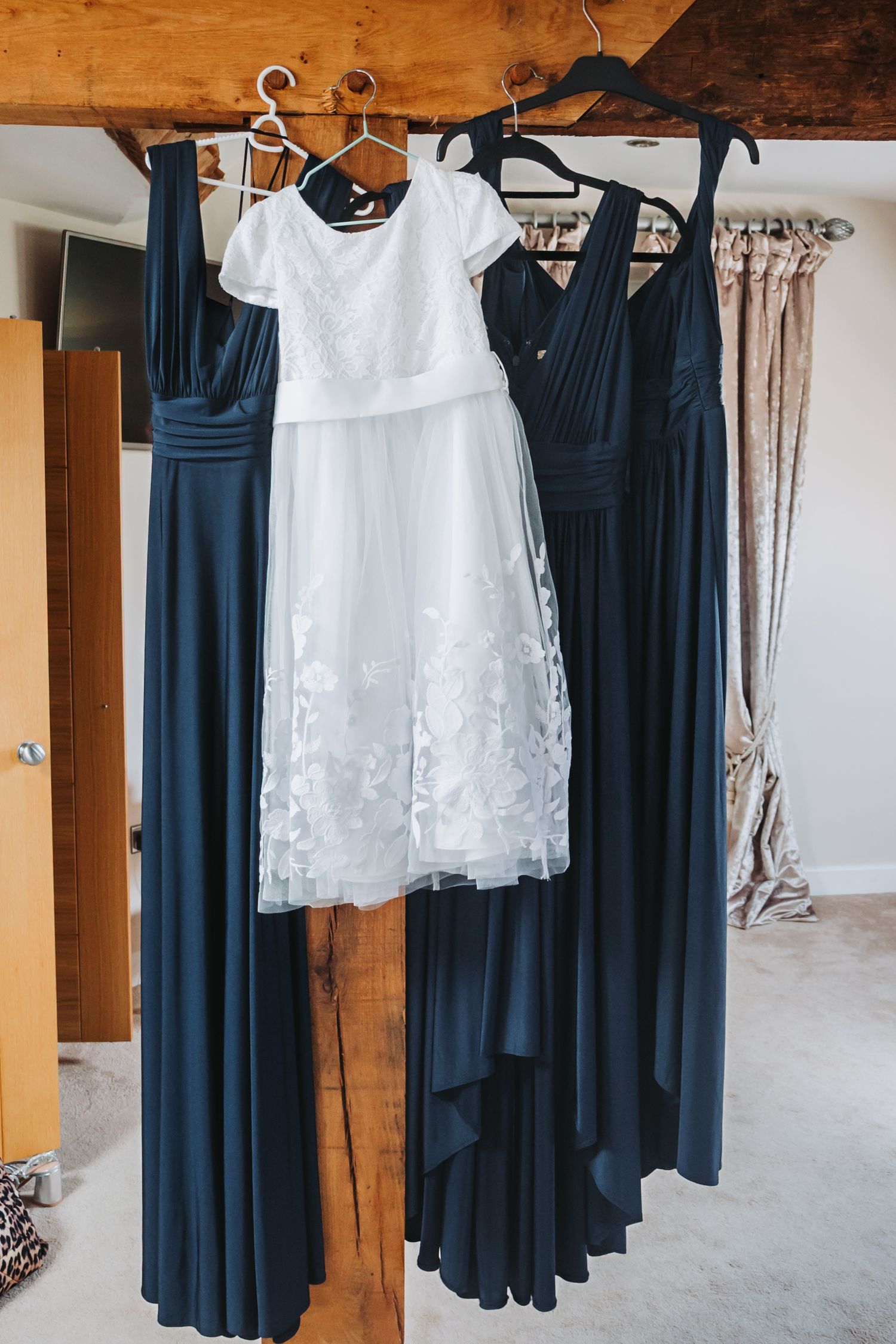 Navy blue bridesmaid dresses and white flower girl dresses hang from wooden beams in a rustic room.