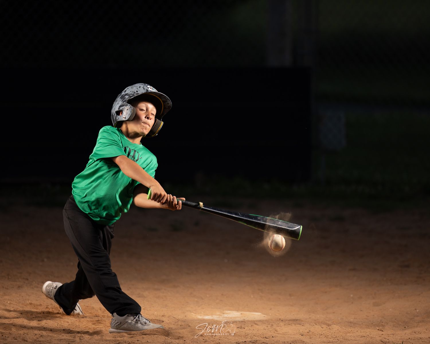 Youth baseball player swinging bat during night game in green jersey.