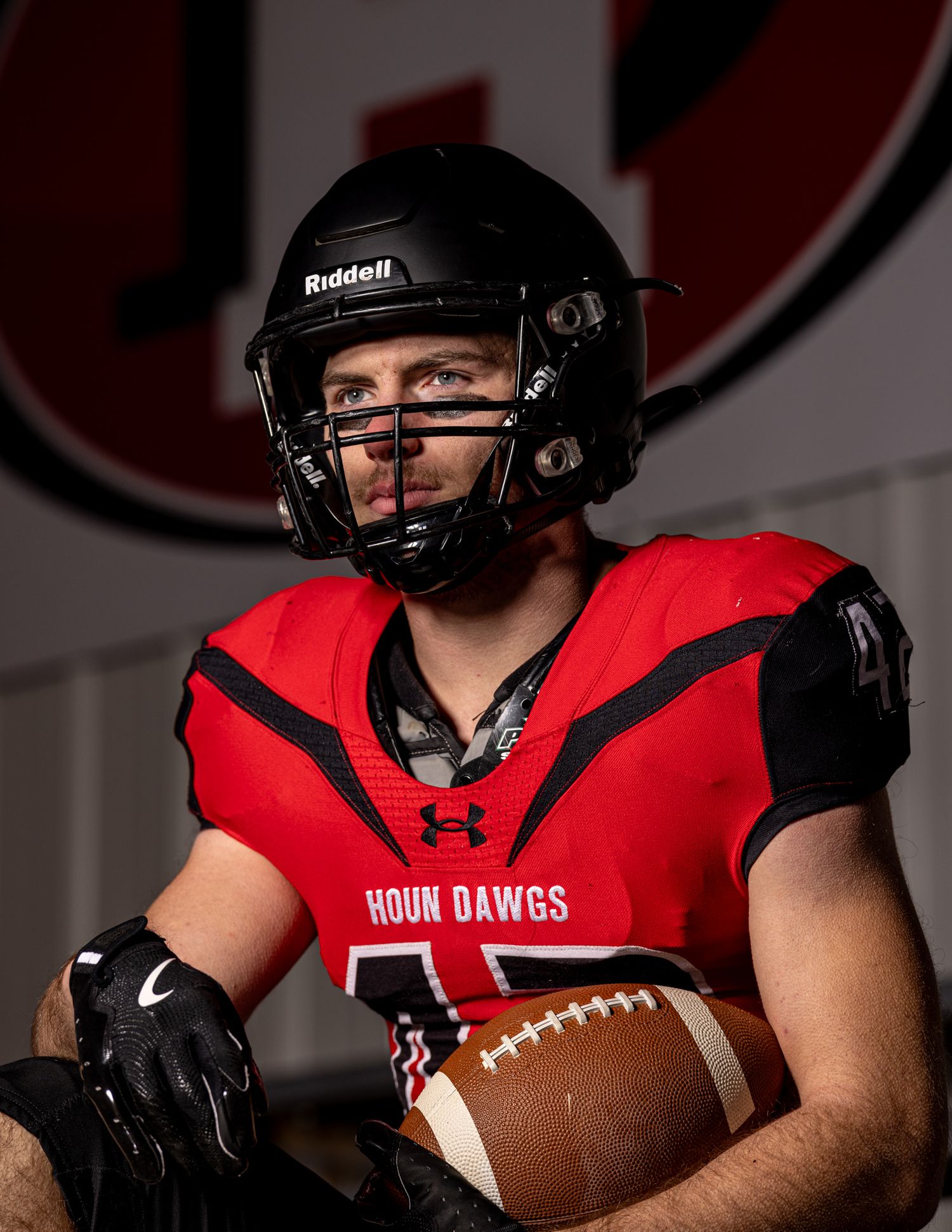 Football player in red uniform holds ball against dark background.