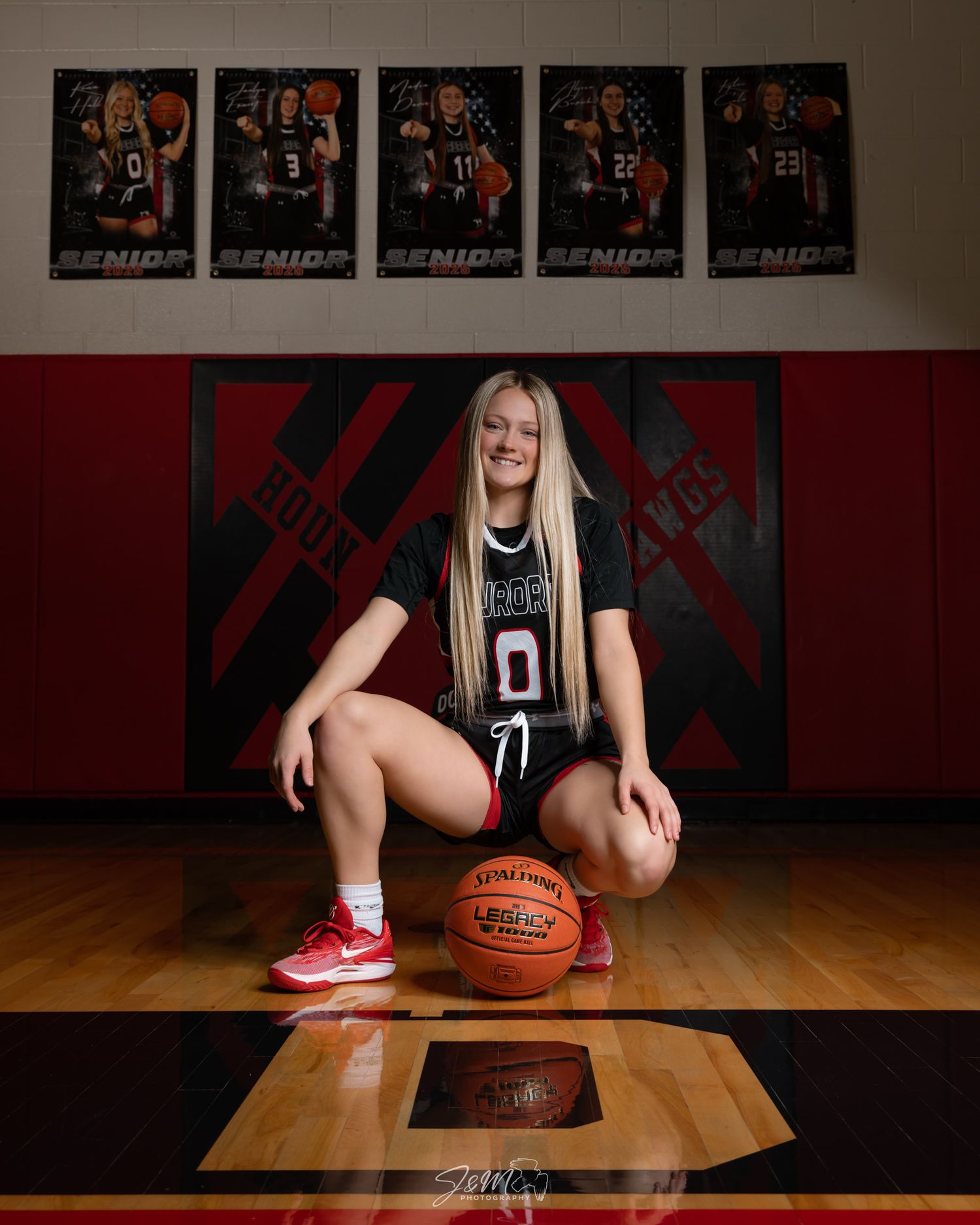 Basketball themed senior portrait with team photos on wall and ball on wooden court.