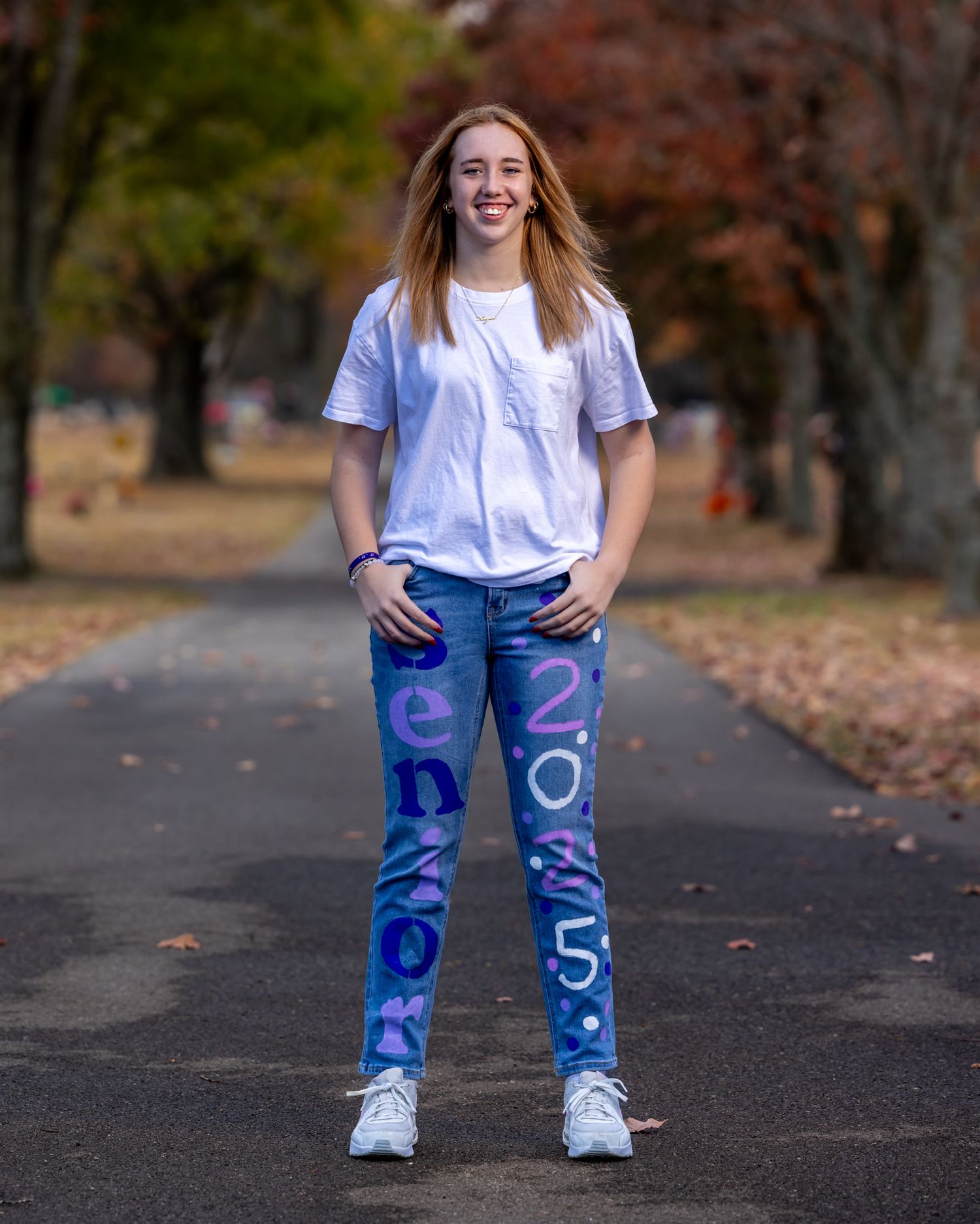 Person standing outdoors wearing white t-shirt and decorated jeans on autumn path.