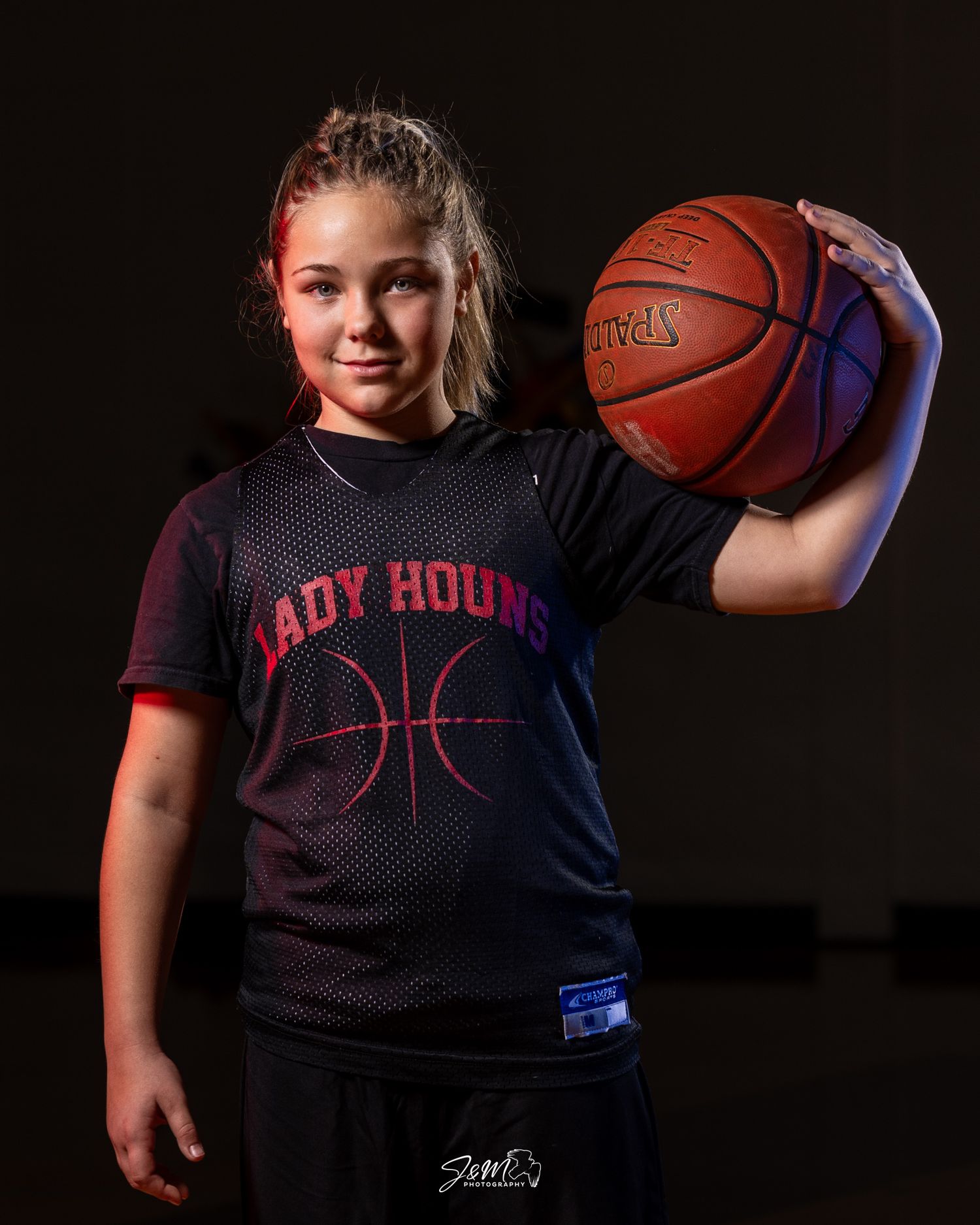 Youth basketball player in Lady Hounds jersey holds basketball against dark backdrop.