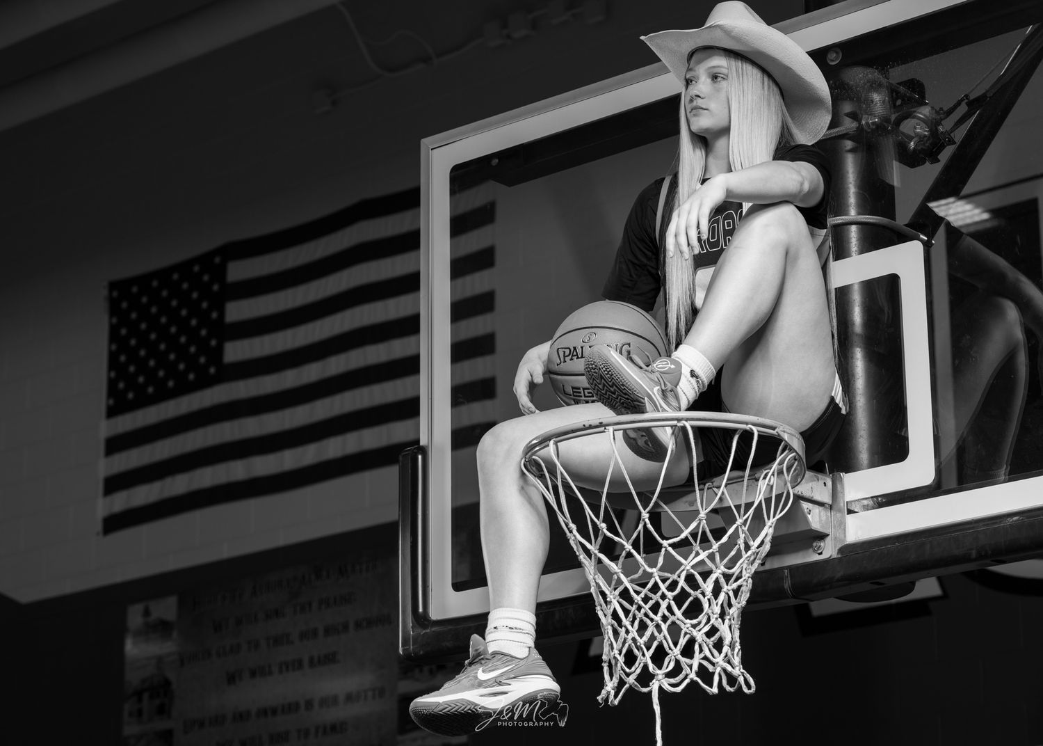 Artistic black and white photo showing someone sitting on a basketball hoop with American flag in background.