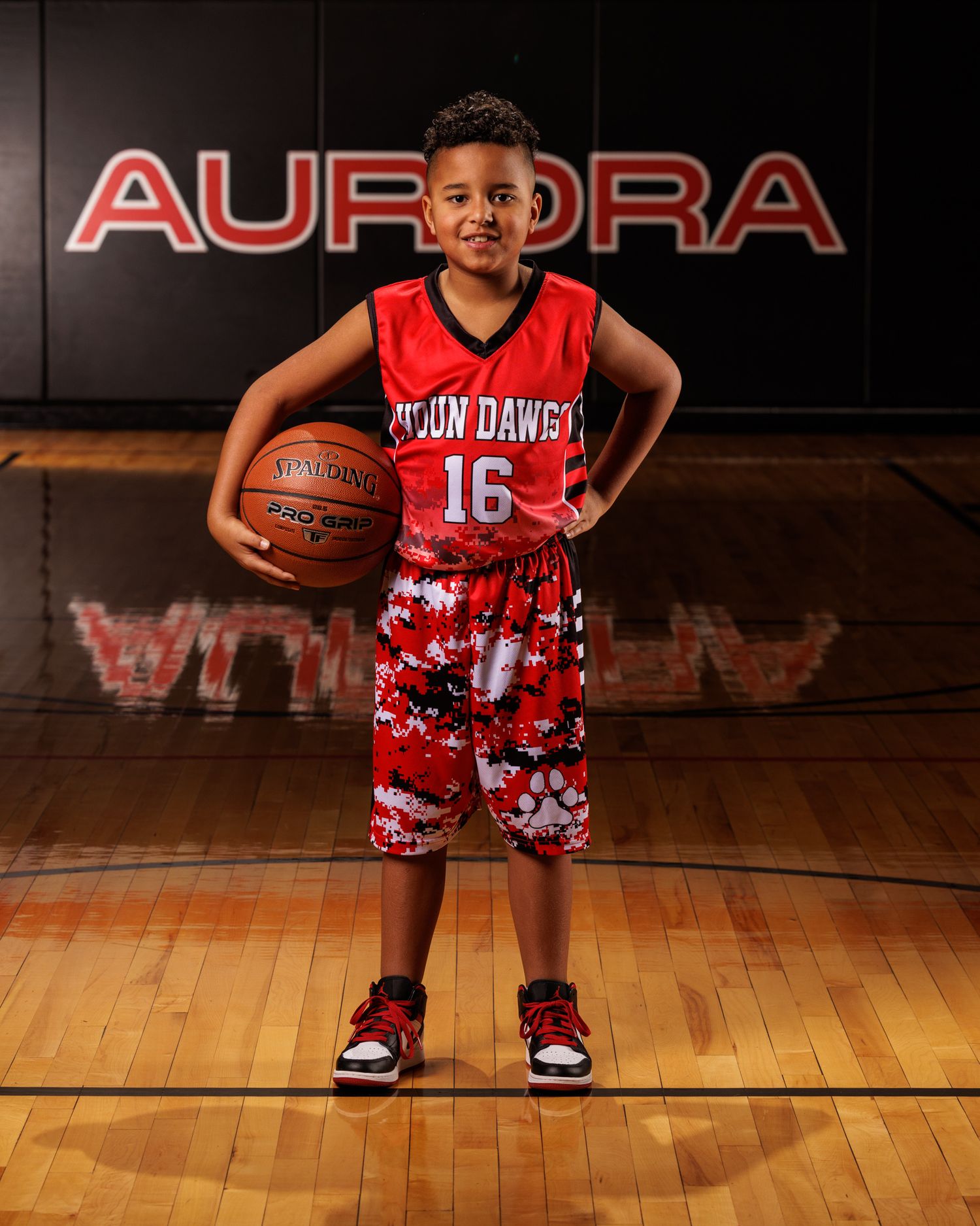 Youth basketball player poses with ball in front of Aurora sign wearing red jersey.