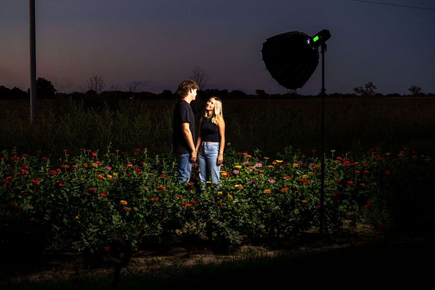 Couple standing in wildflower field at dusk with dramatic lighting and photography equipment visible.