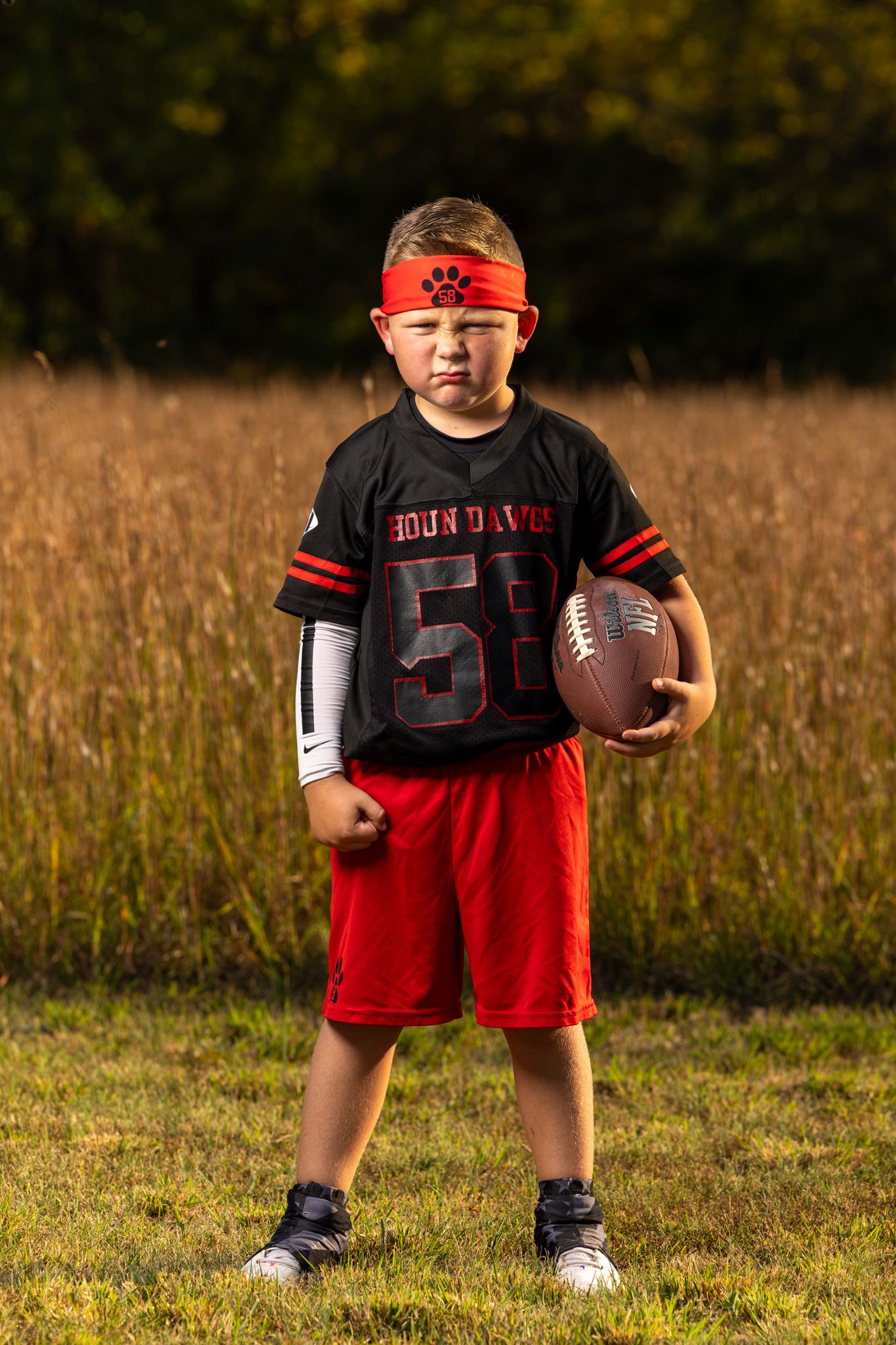 Young football player poses in black jersey and red shorts while holding football in grassy field.
