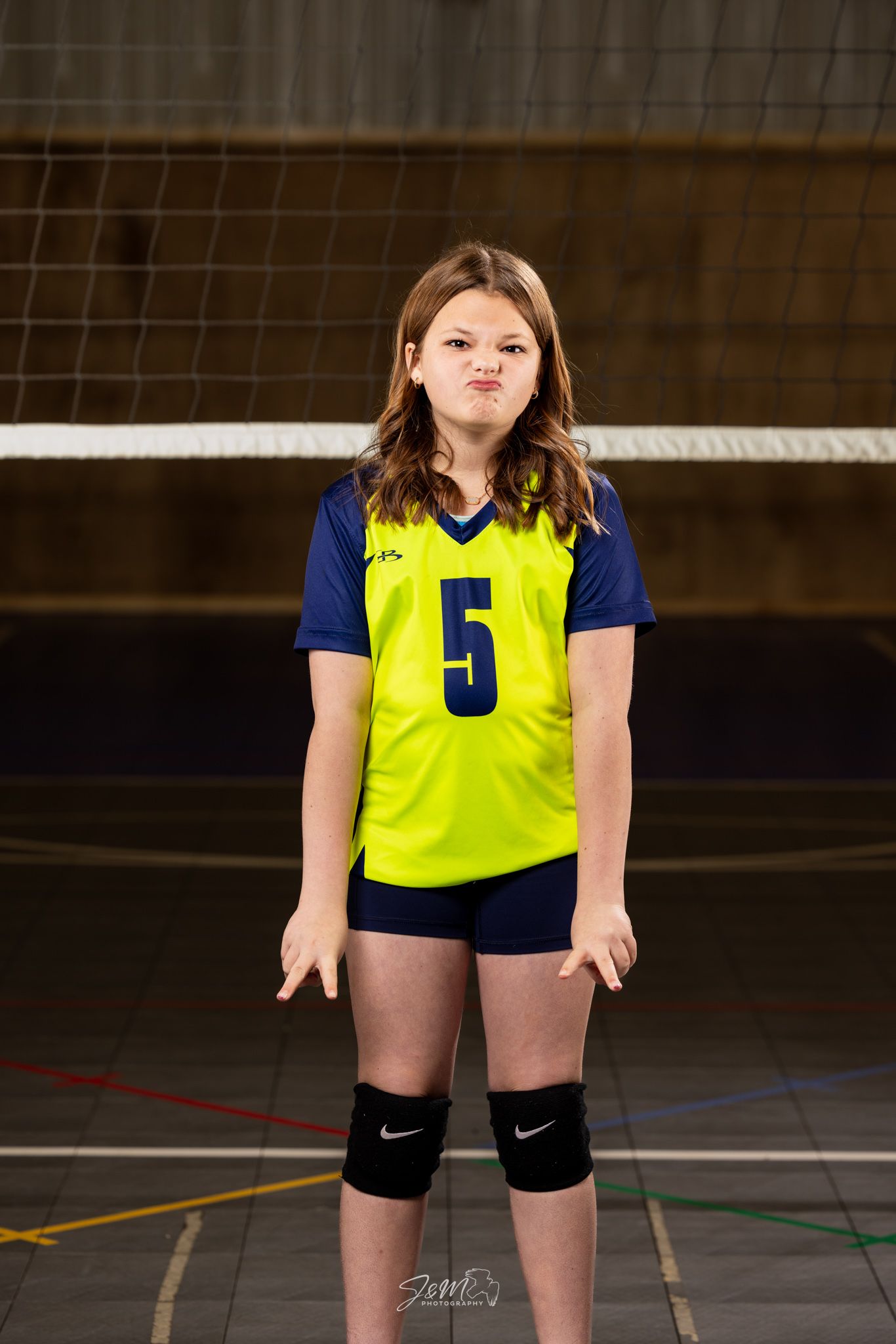 Volleyball player wearing yellow jersey number 5 and navy shorts stands in front of net with knee pads.