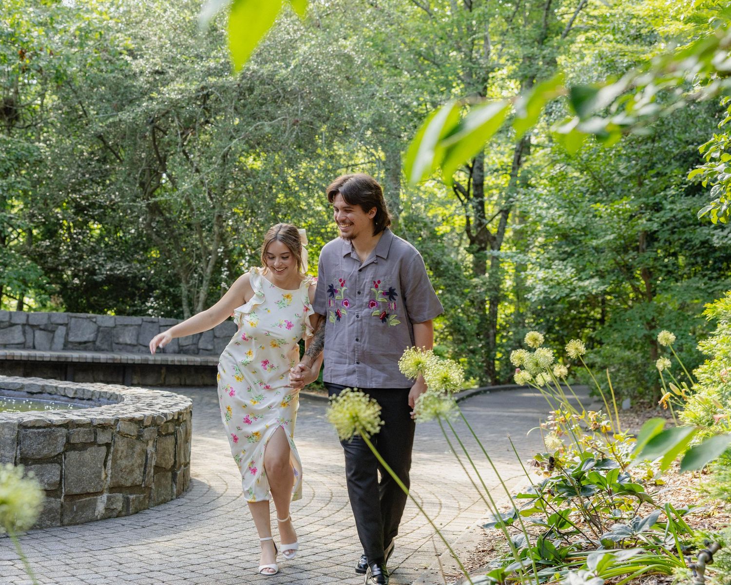 A couple strolls together along a winding stone path surrounded by lush green foliage on a sunny summer day.