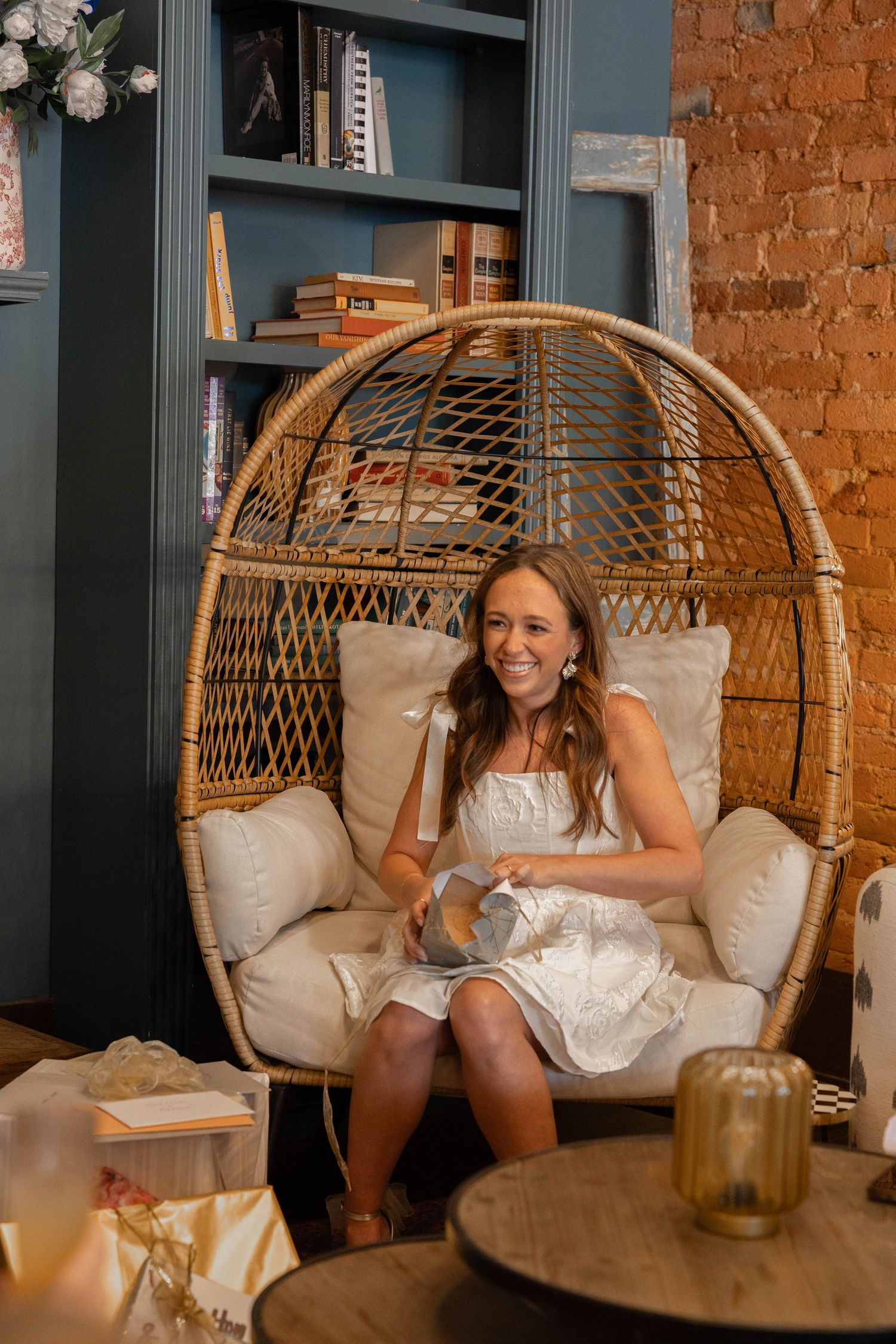 A person sits comfortably in a hanging rattan egg chair with plush cushions inside a stylish brick interior space.