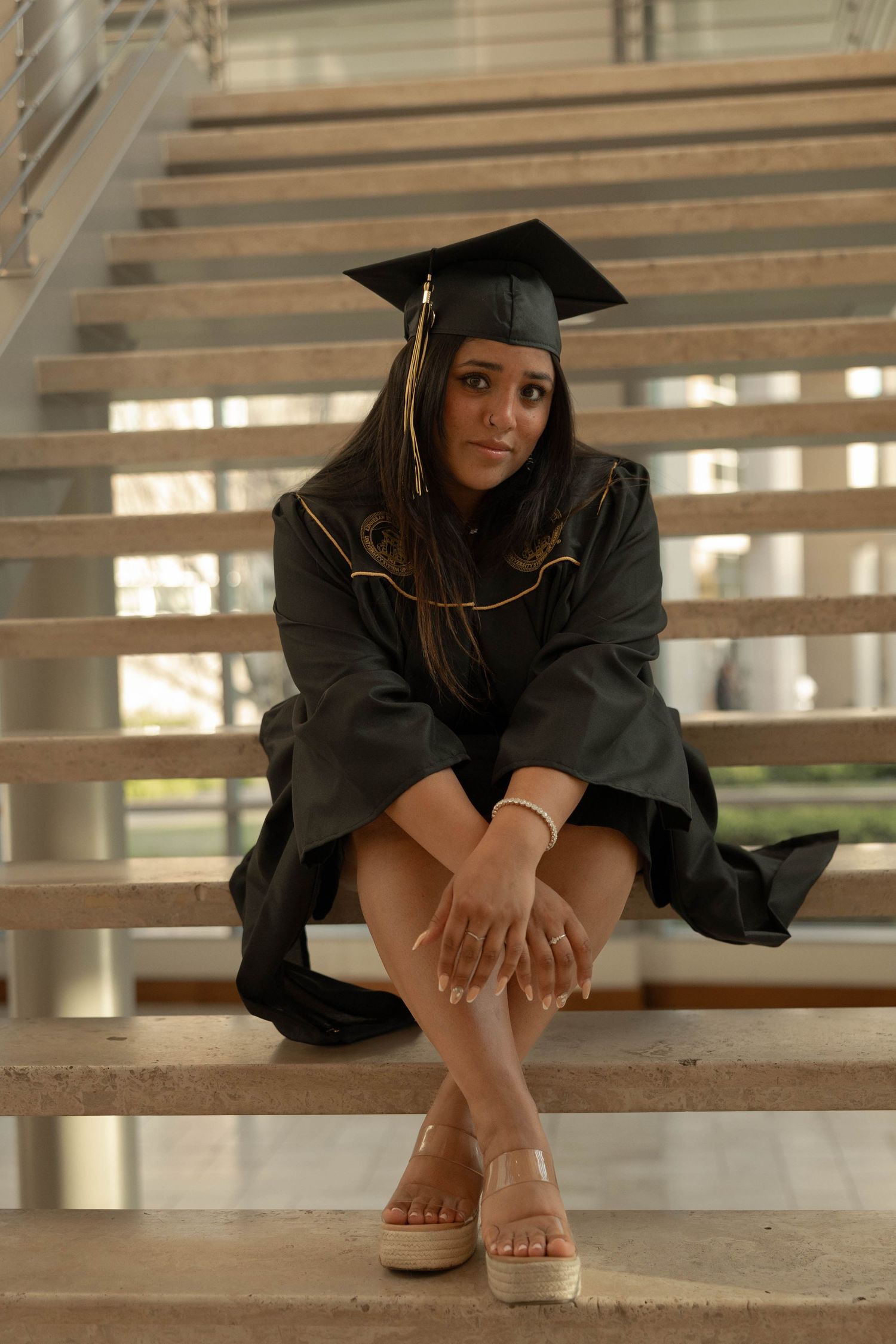 Kennesaw State University Graduate in cap and gown sitting cross-legged on concrete stairs indoors.