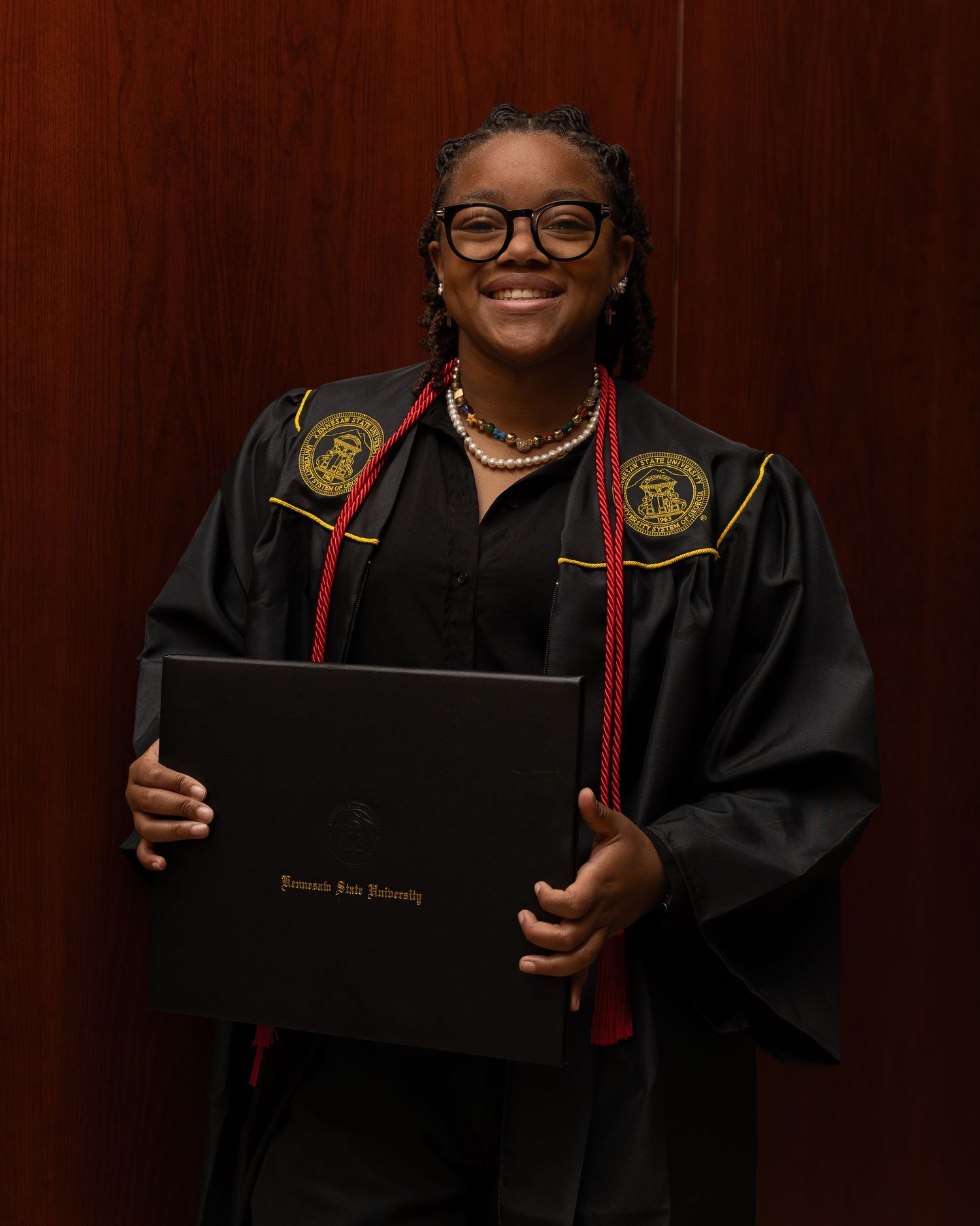 Graduate in academic regalia poses with diploma against dark wood paneling in professional portrait series.