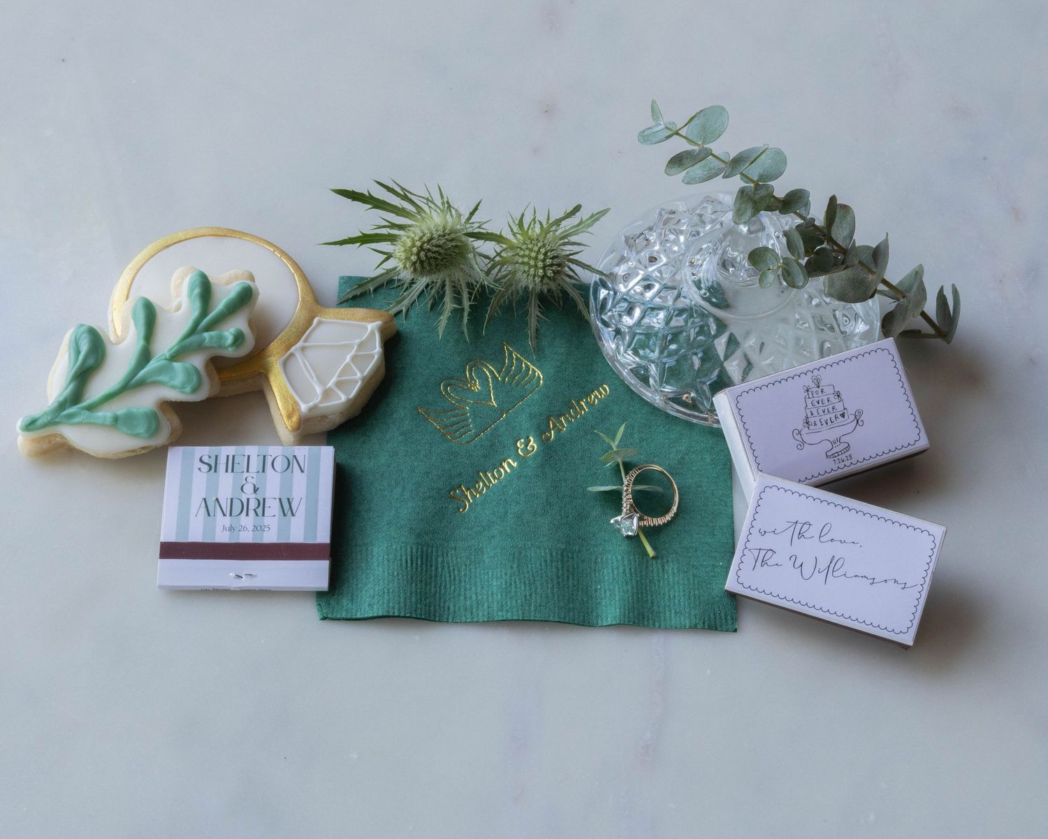 A collection of wedding-themed items including a green fabric pouch, sugar cookie, gift tags, and greenery accents.