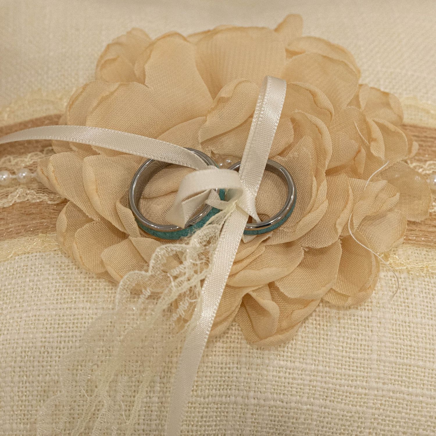 Wedding rings tied with white ribbon to a beige fabric flower on lace and burlap textured background.