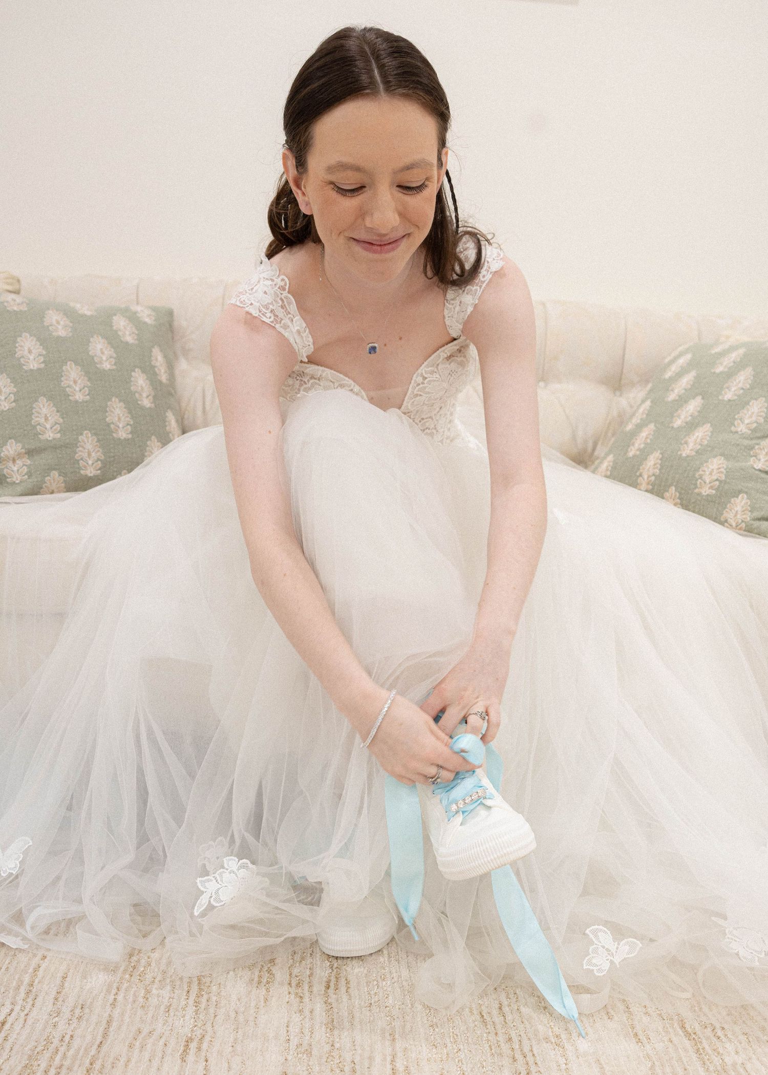 A bride in a white dress sits on floor arranging pale blue wedding shoes with tulle surrounding her.