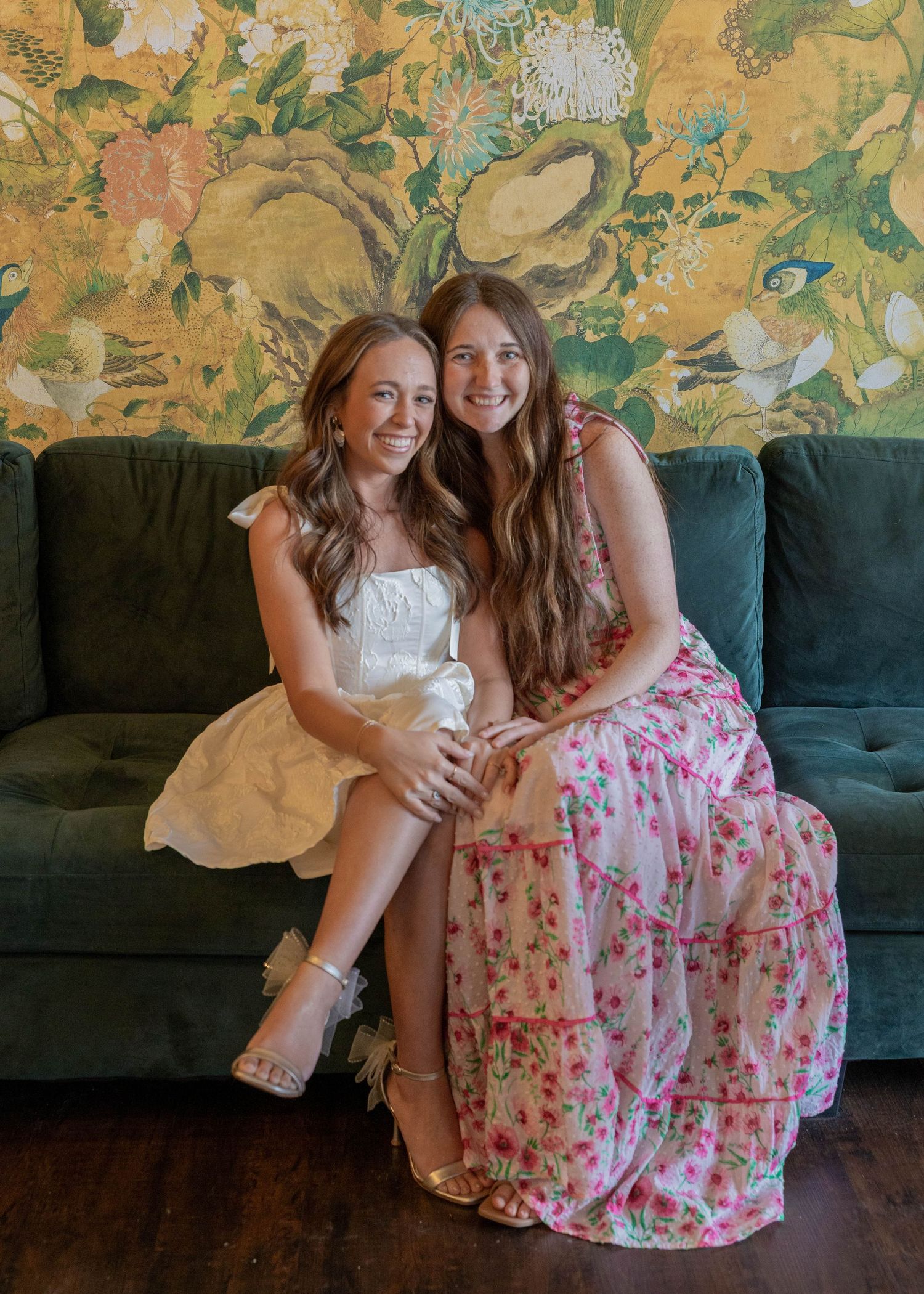 Two people wearing floral dress and white dress sit on green couch against a gold floral wallpaper background.