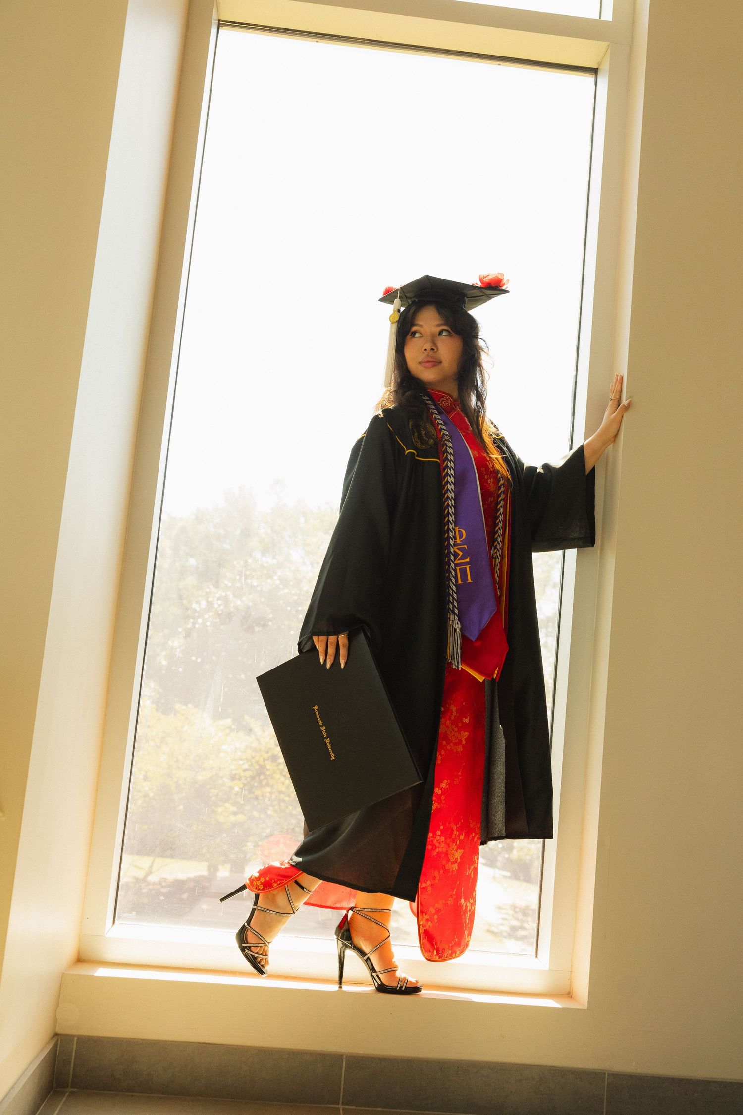 Graduate in black cap and gown poses near bright windows during graduation photo session.