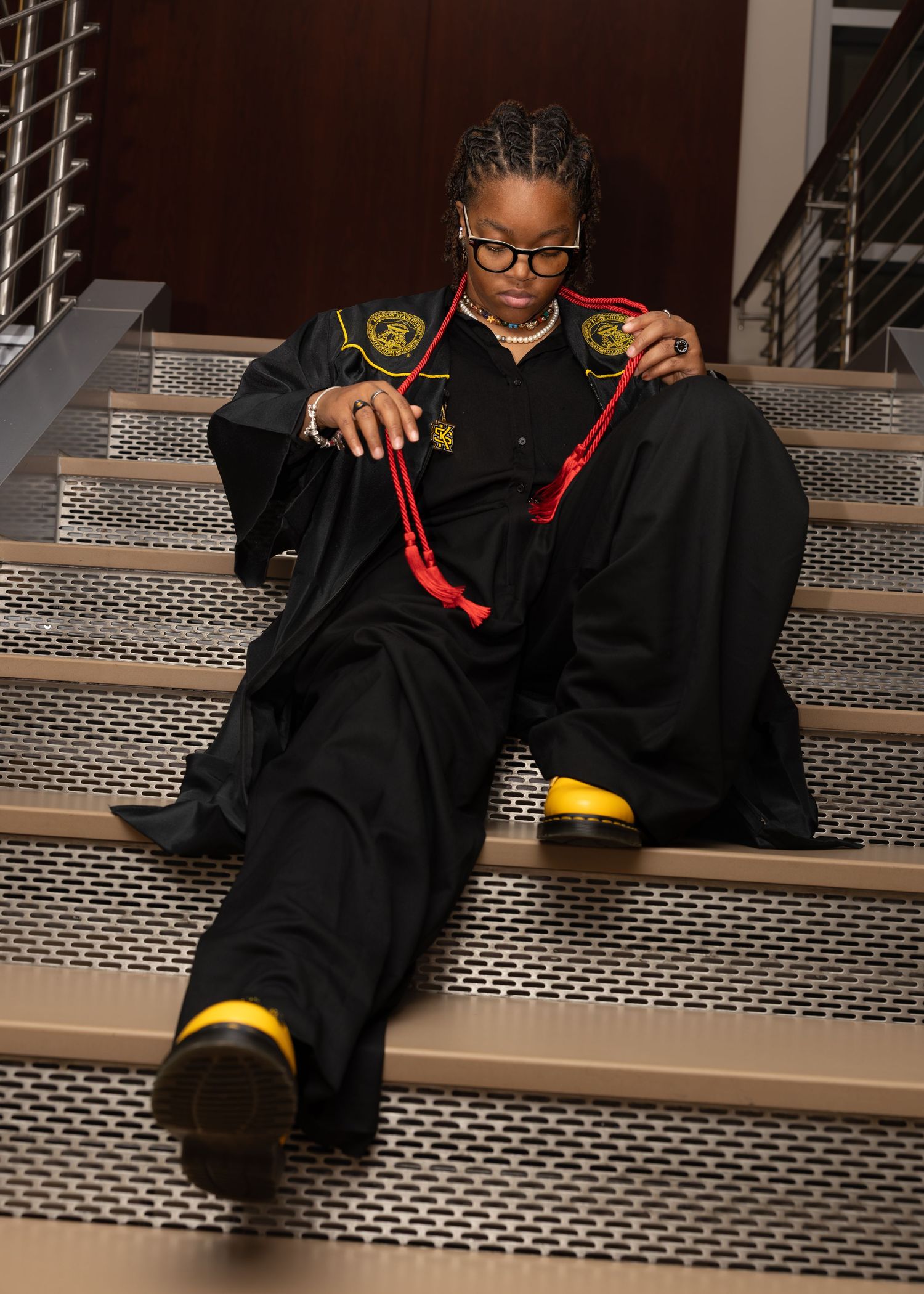 Person in black graduation gown and glasses sits on metal stairs while looking down at their feet.