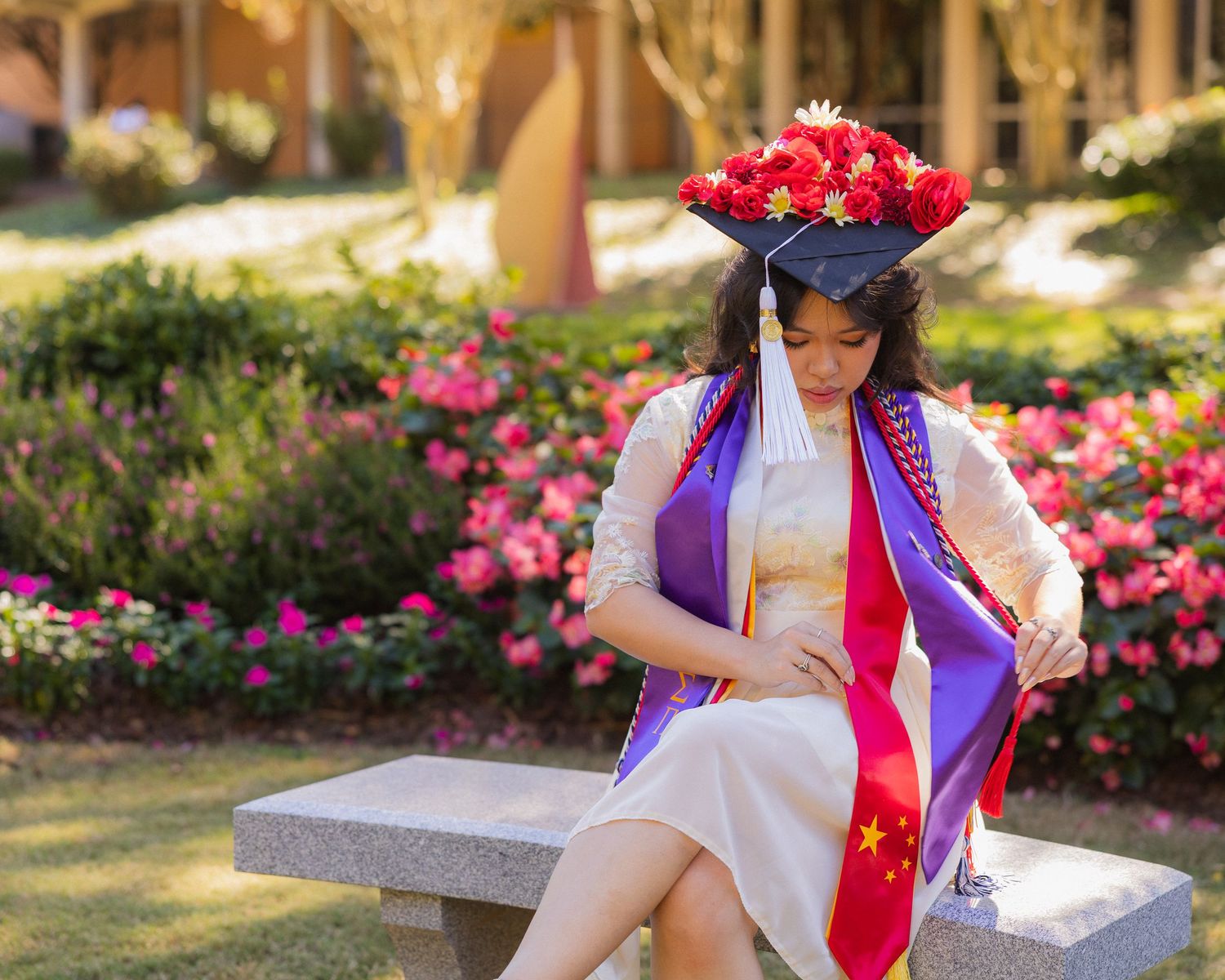A person in a blue and white outfit sits on a stone bench surrounded by blooming pink roses in a garden setting.
