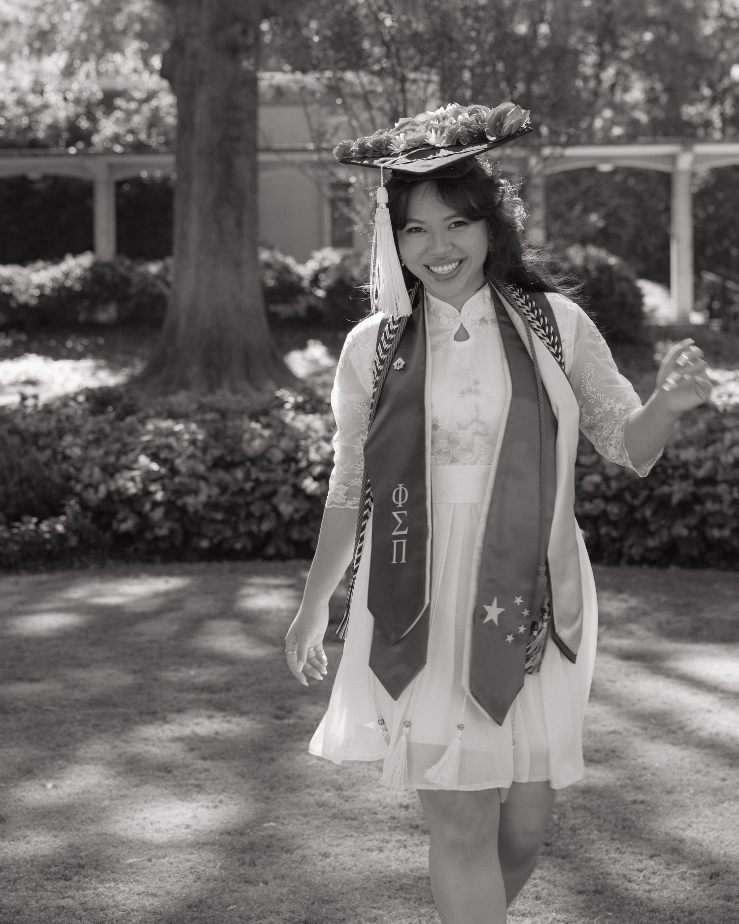 A graduate wearing a decorated cap and white dress with graduation stoles poses outdoors in a black and white photo.