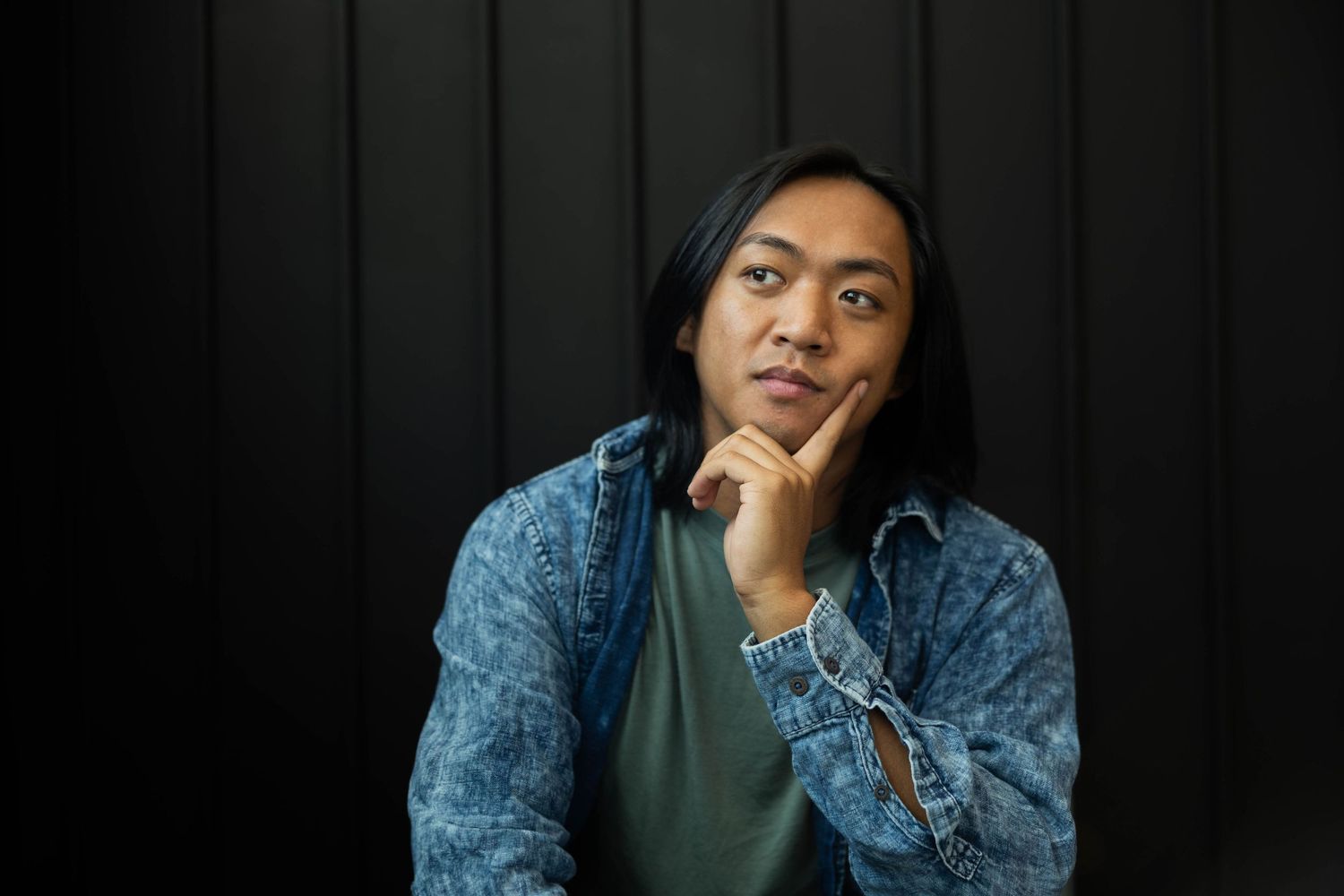 Person in denim jacket poses against dark background in casual portrait series.