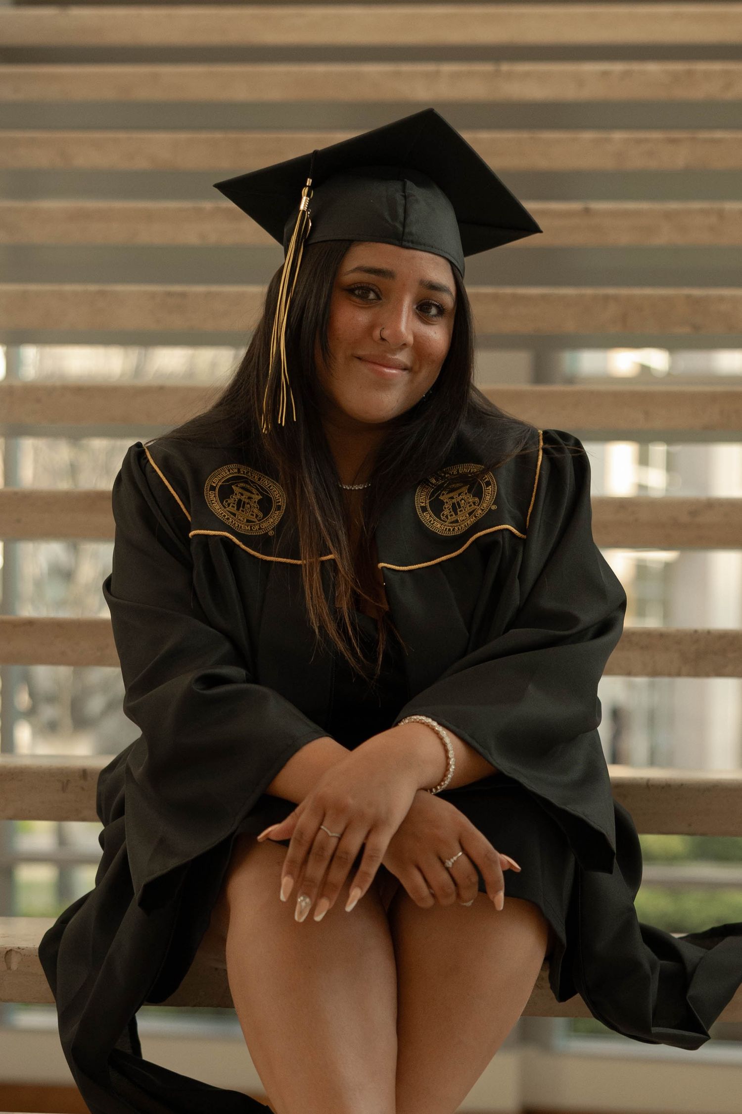 A graduate wearing traditional black cap and gown with gold trim poses on wooden steps.