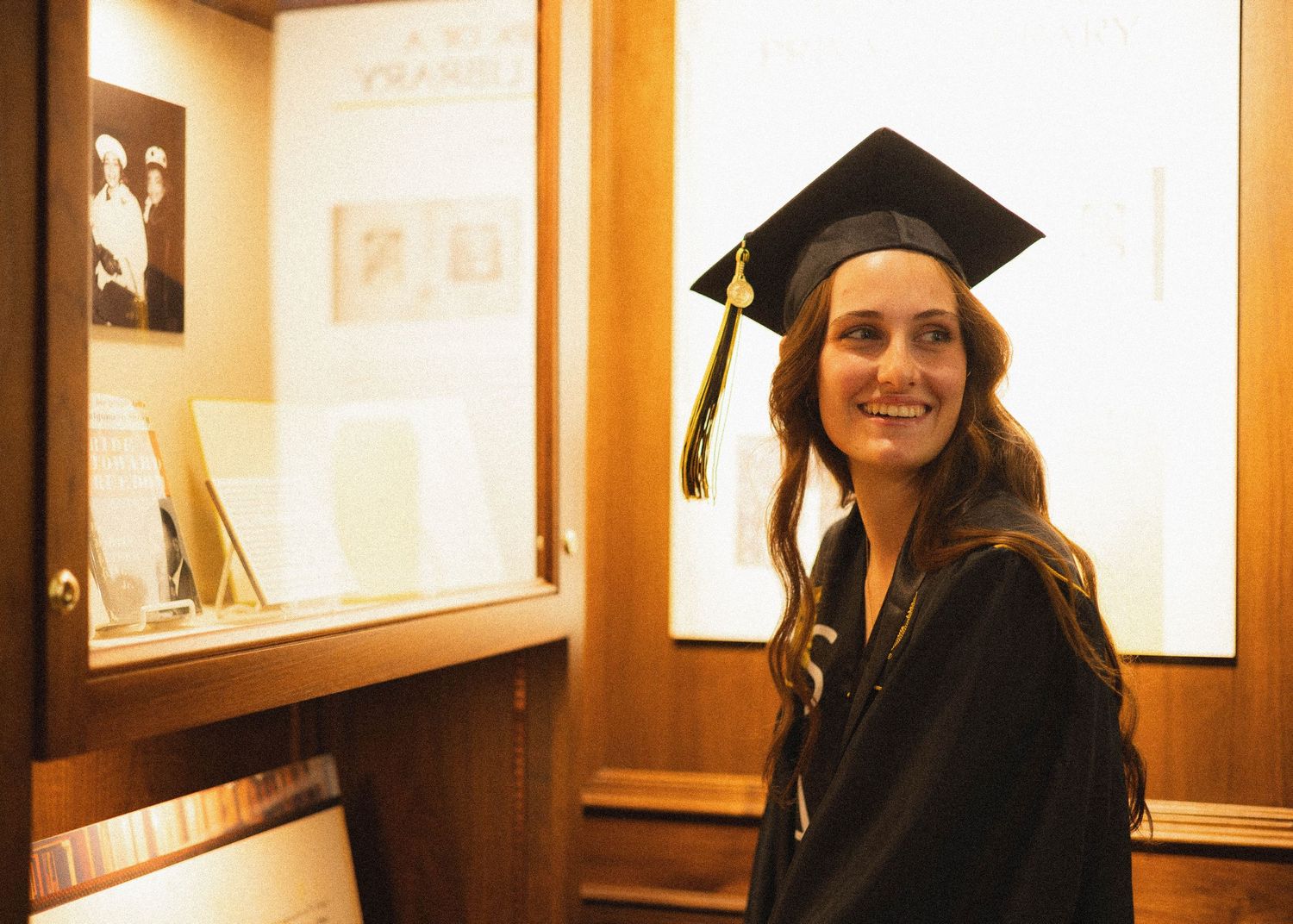 Graduate wearing academic regalia stands in warmly lit display room.