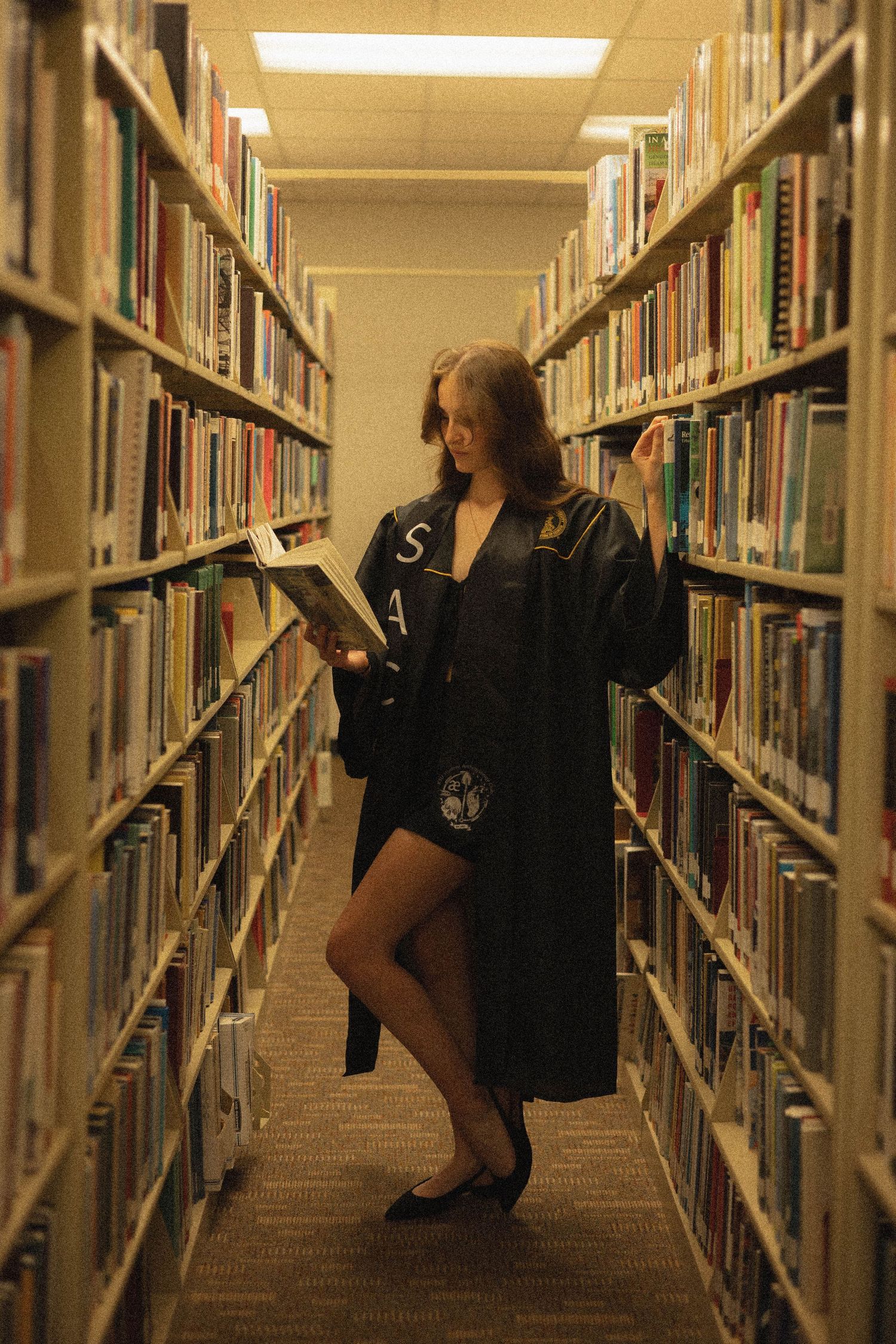 Figure in black attire reading while standing in a library aisle.