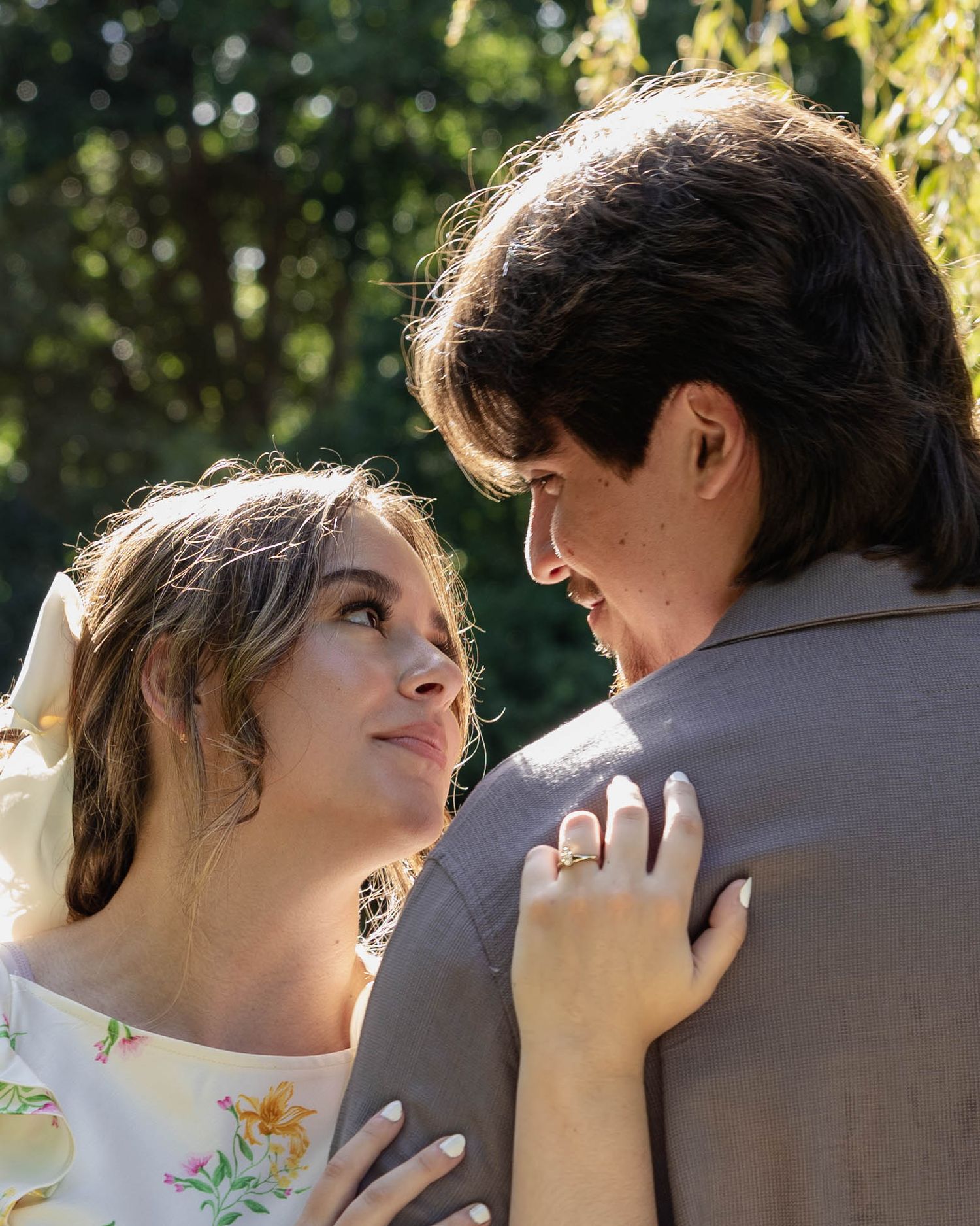A tender romantic moment between a couple standing close together outdoors with sunlit foliage in the background.