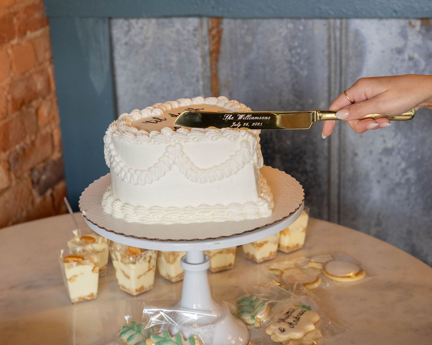 Hands decorating and assembling a rustic white wedding cake on a pedestal stand in a bakery kitchen.
