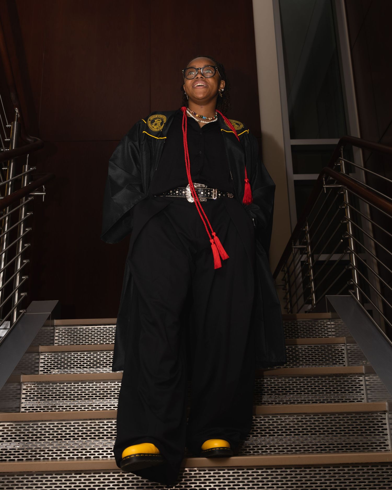 Graduate wearing black cap and gown poses on metal staircase in low light conditions.