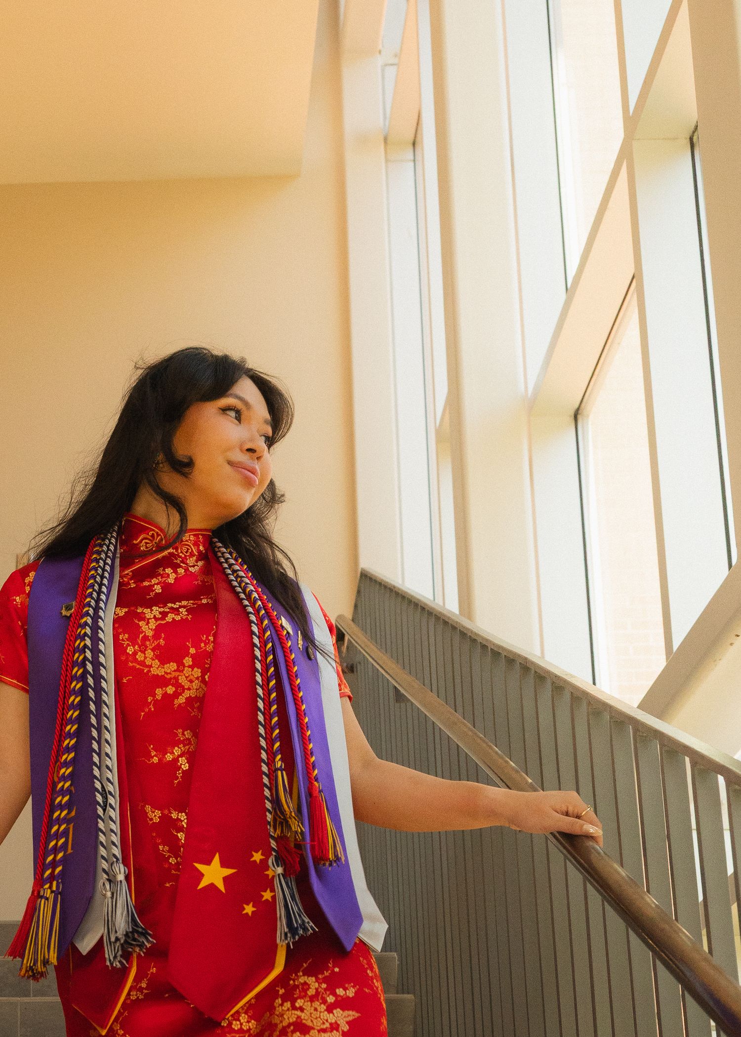 Person in traditional red Chinese qipao dress with floral pattern and blue scarf poses playfully near a stairway railing.