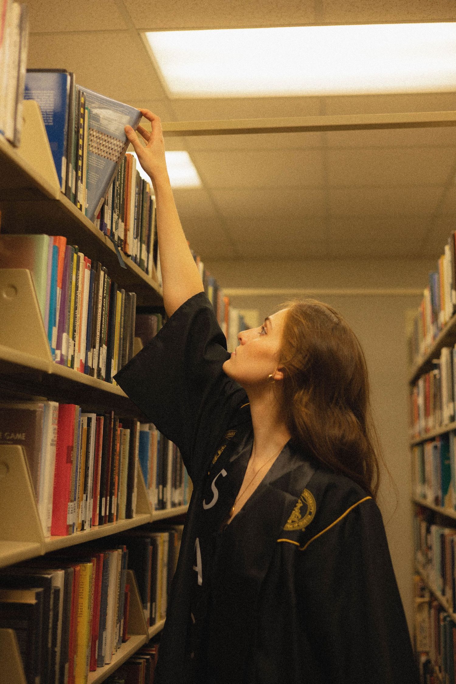 A student reaches up to get a book from a tall library shelf while wearing a dark sweater.