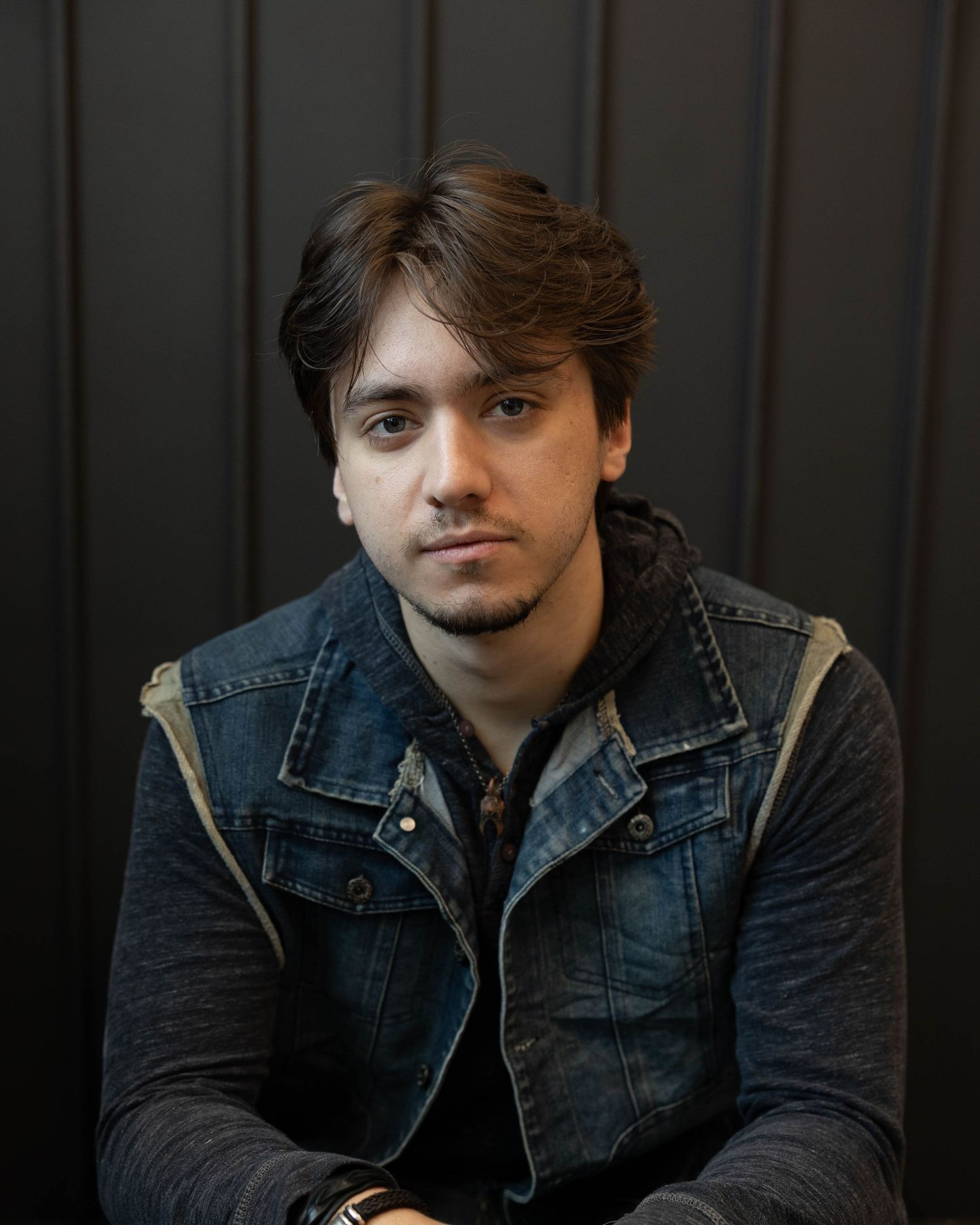 Portrait of a young person in a denim vest with dark sleeves against a dark background.