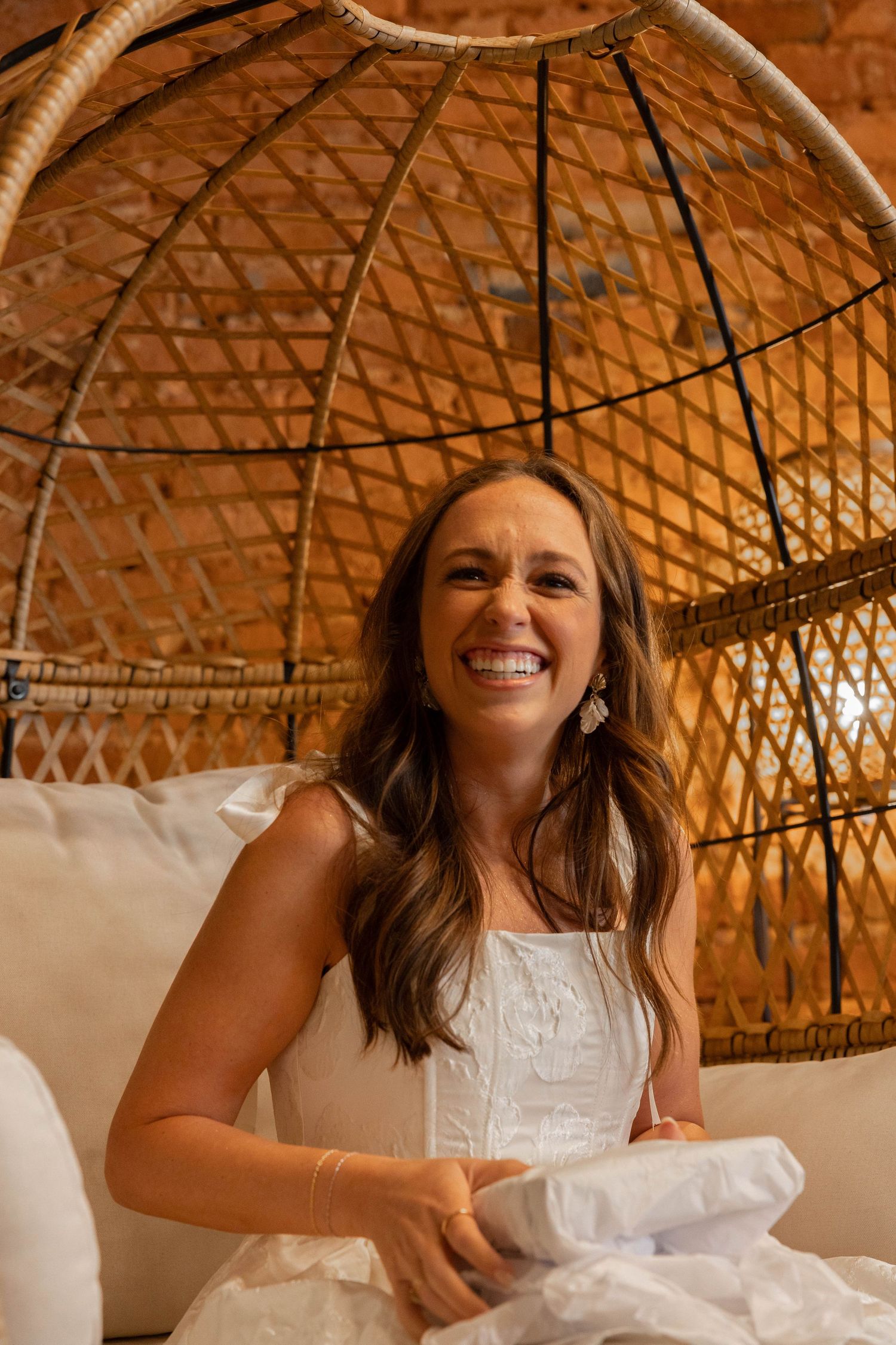 Joyful person in white dress sitting on wicker chair with decorative peacock-style backing in warm lighting.