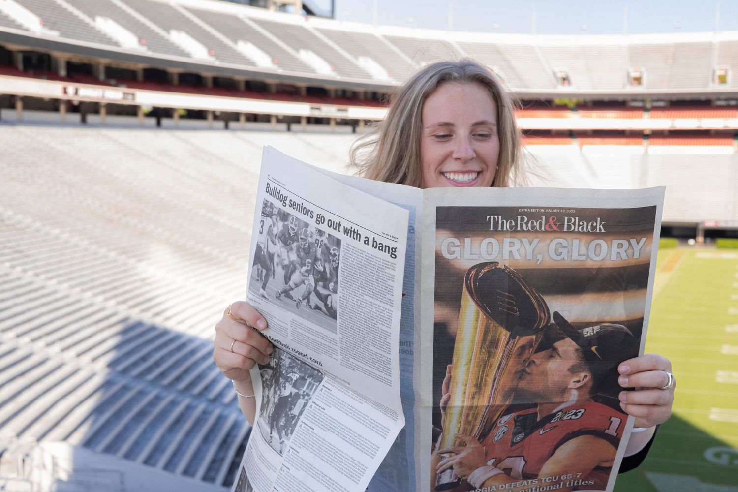University of Georgia Graduate holding newspapers while sitting in empty stadium bleachers.